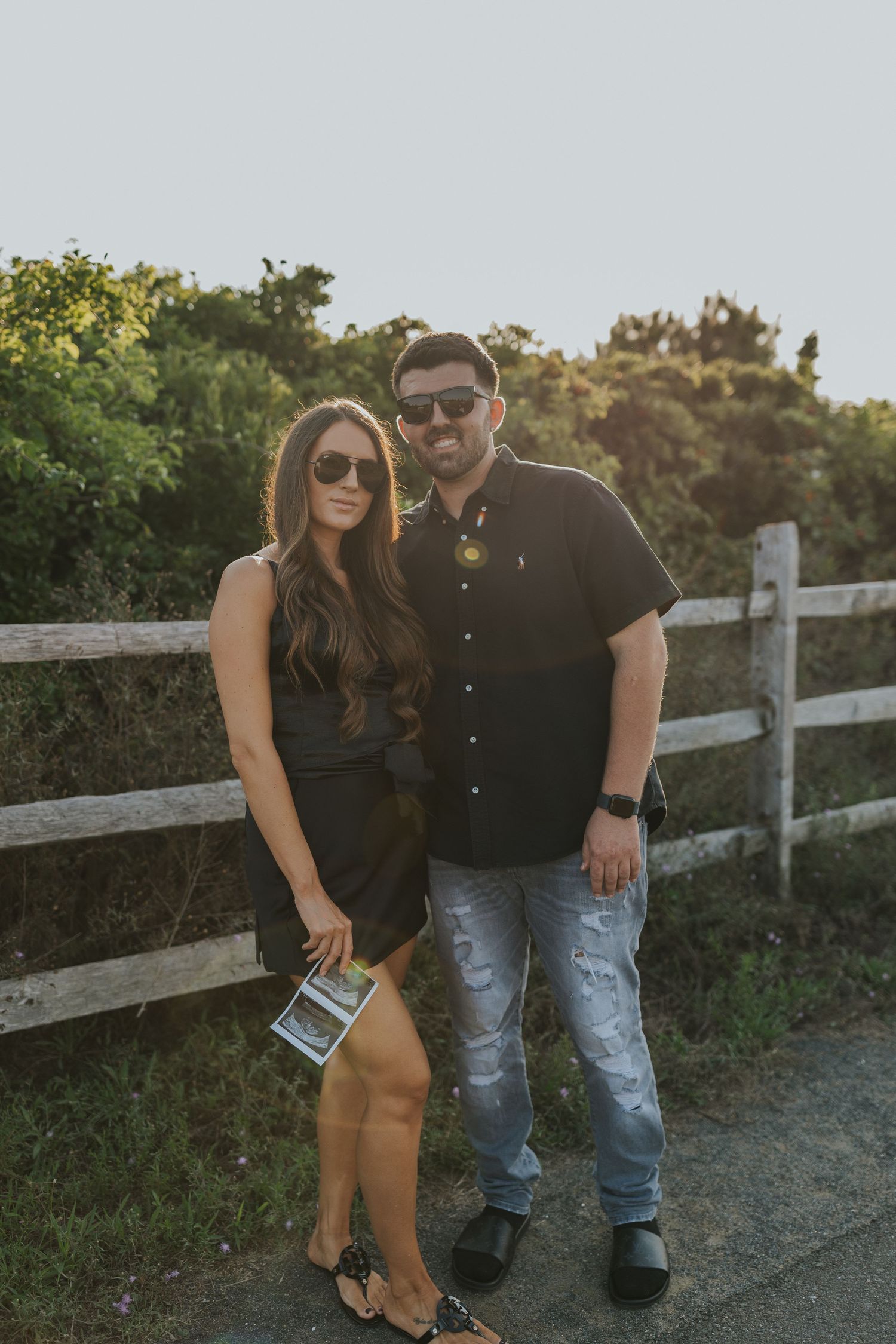 Stylish couple wearing sunglasses standing by white fence at golden hour, casual outdoor portrait.