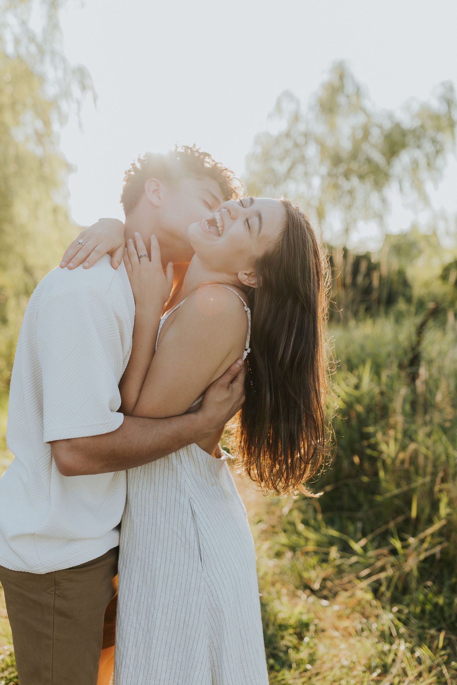 Couple sharing a joyful kiss outdoors in golden sunlight surrounded by lush green nature.