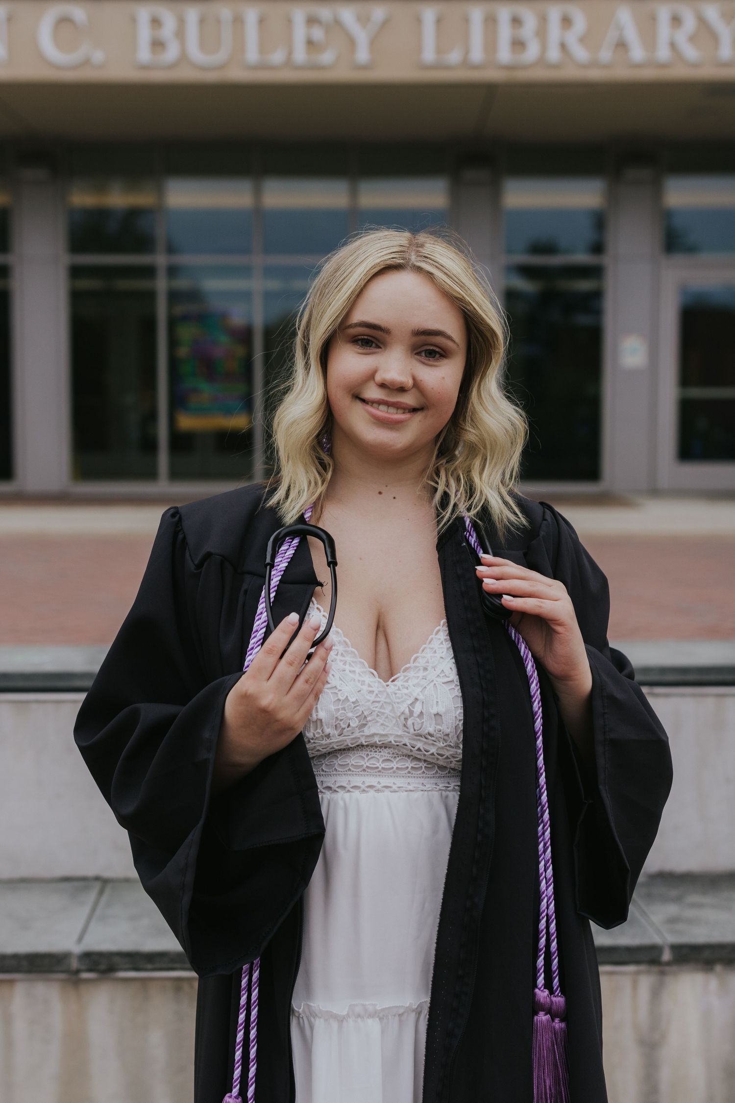Smiling graduate in black gown and purple honor cord holding cap outside Buley Library.