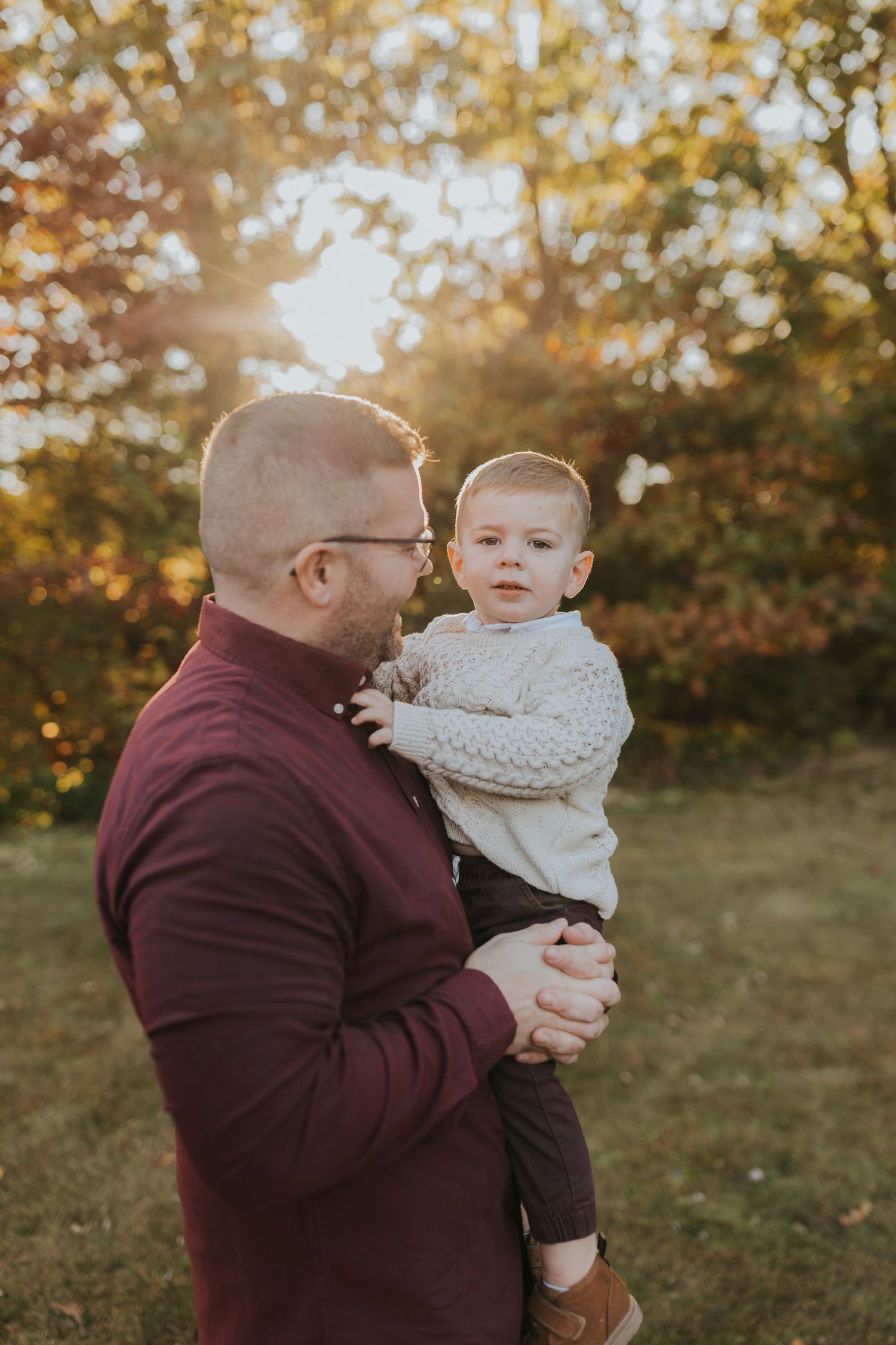 Father holding toddler son in cream knit sweater during golden hour fall family photo session outdoors.