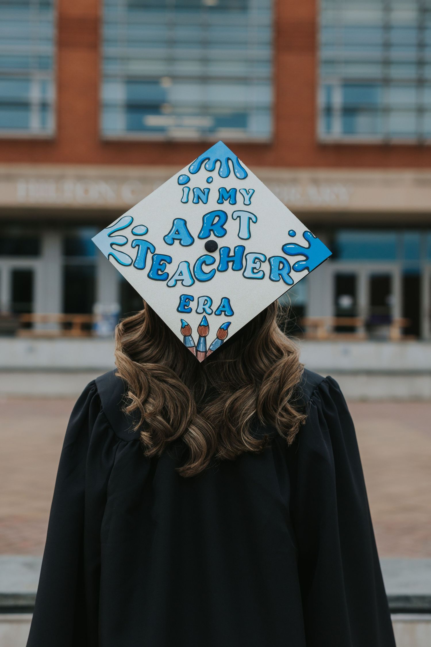 Graduate displays decorated mortarboard reading In My Art Teacher Era outside university library.