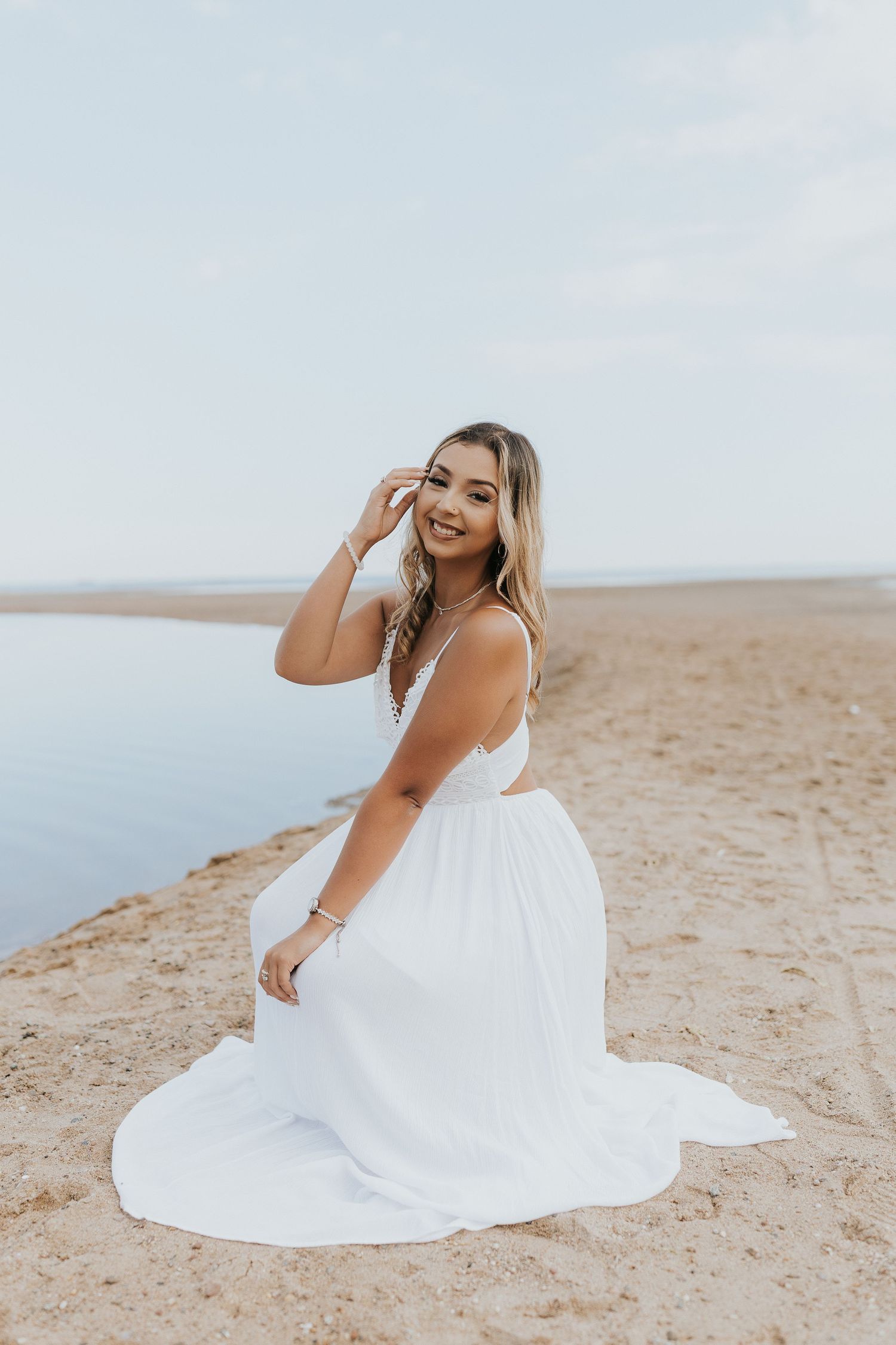 Smiling young woman in flowing white dress sits on sandy beach shore beside calm blue water.