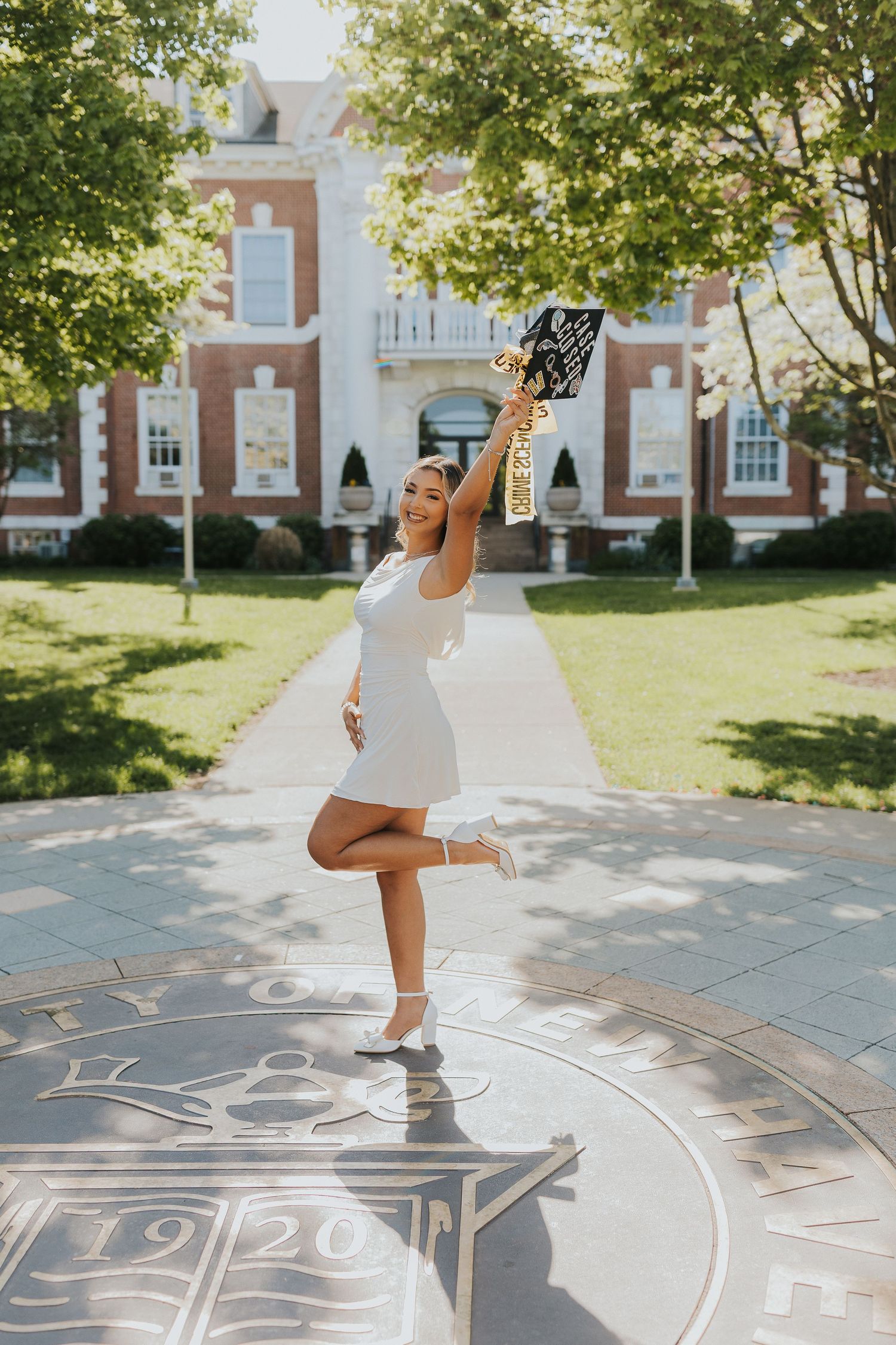 Joyful female graduate in white dress raising cap on university campus seal outdoors.