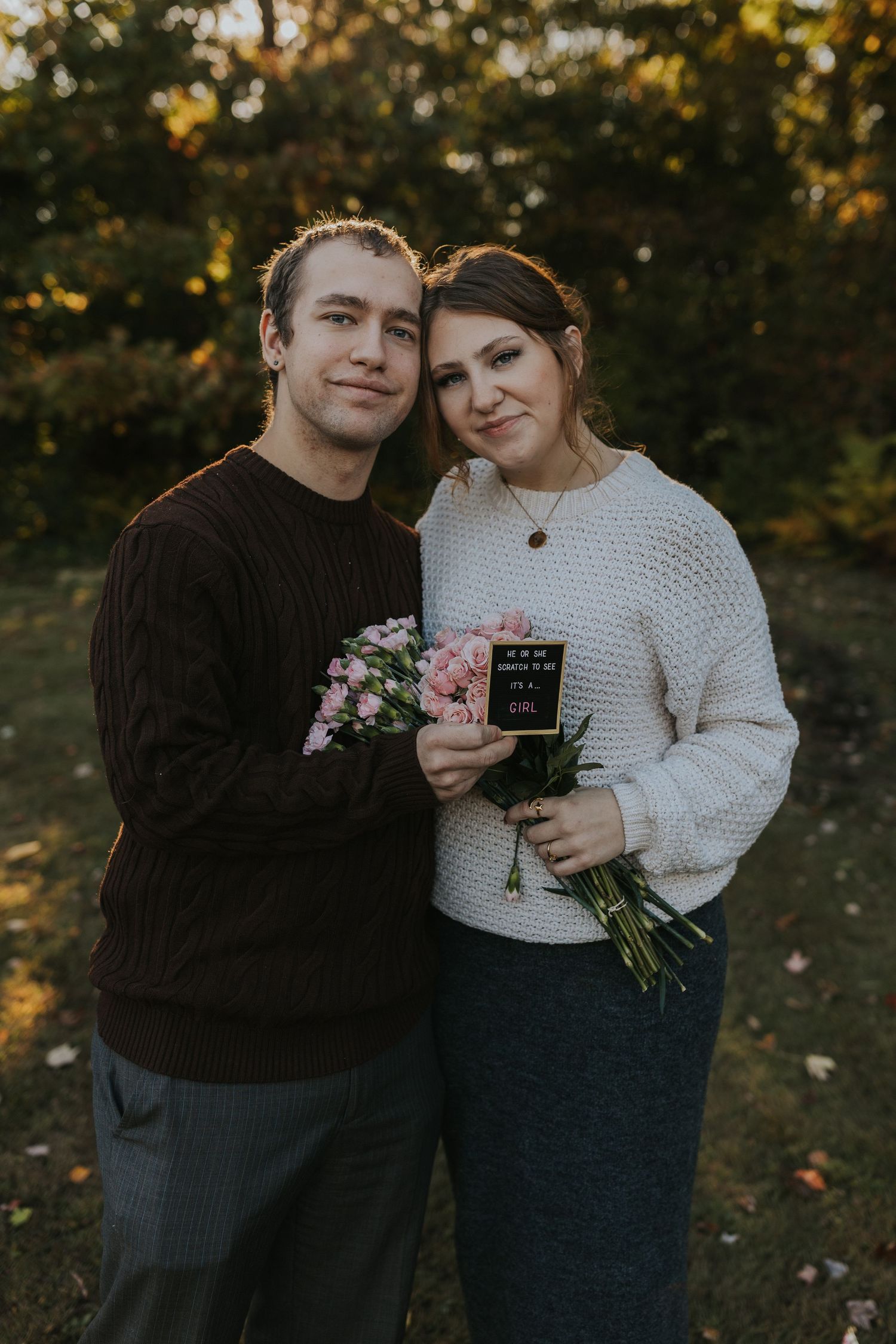 Couple holding pink flowers and small sign during outdoor fall pregnancy announcement photo session at sunset.