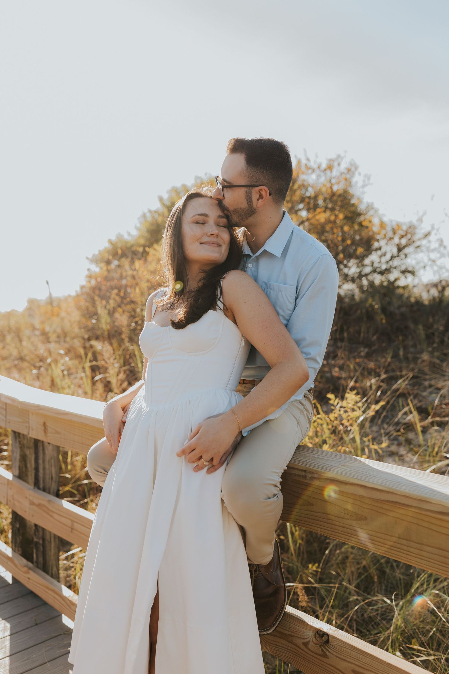 Couple embracing on wooden boardwalk at sunset, man kissing woman in white dress tenderly.