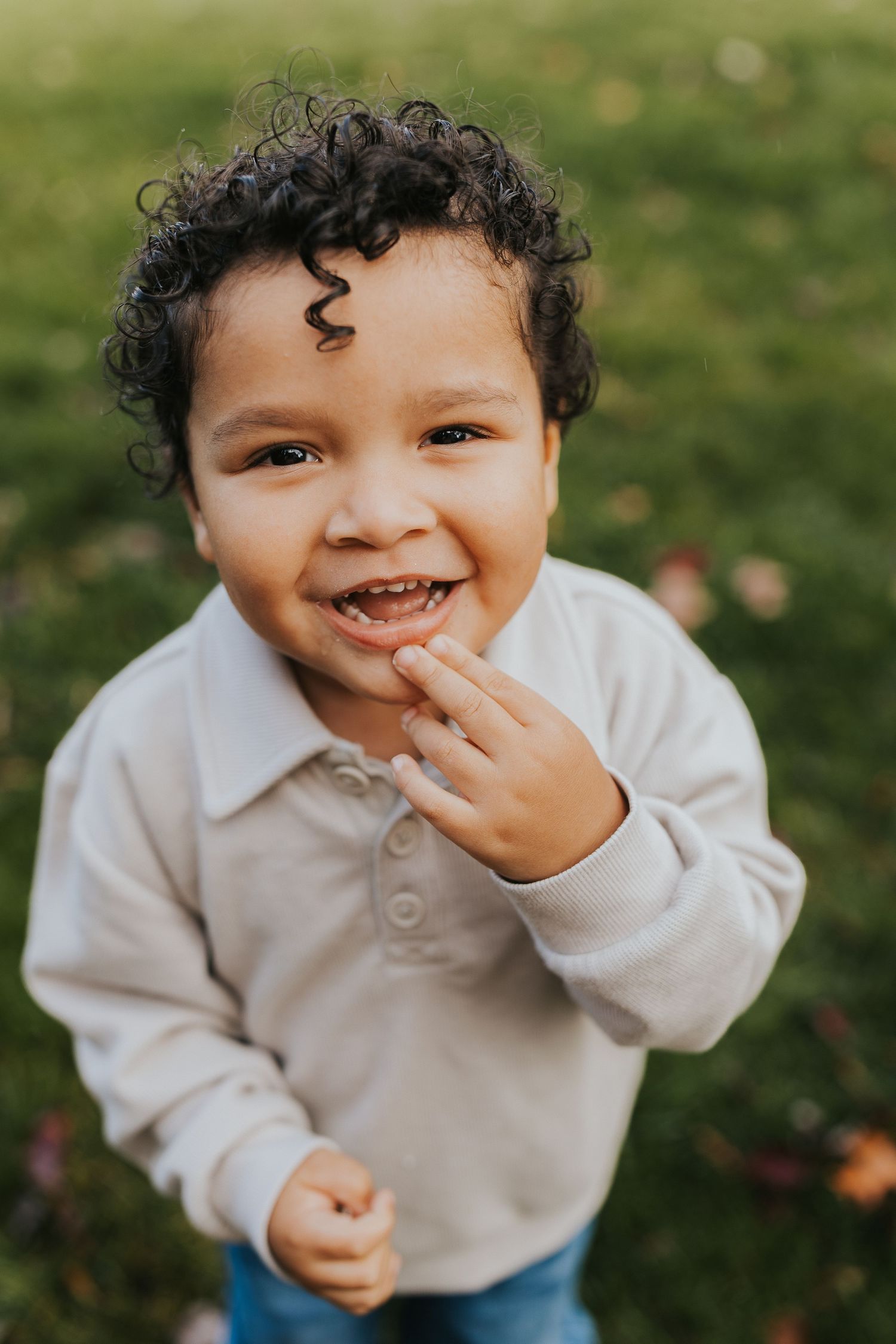 Smiling toddler boy with curly hair wearing a cream polo sweater outdoors in a green park.