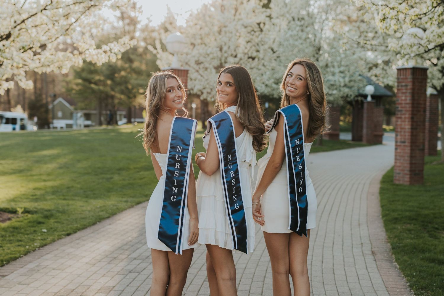 Three female graduates in white dresses wearing blue graduation stoles pose on a blooming tree-lined campus walkway.