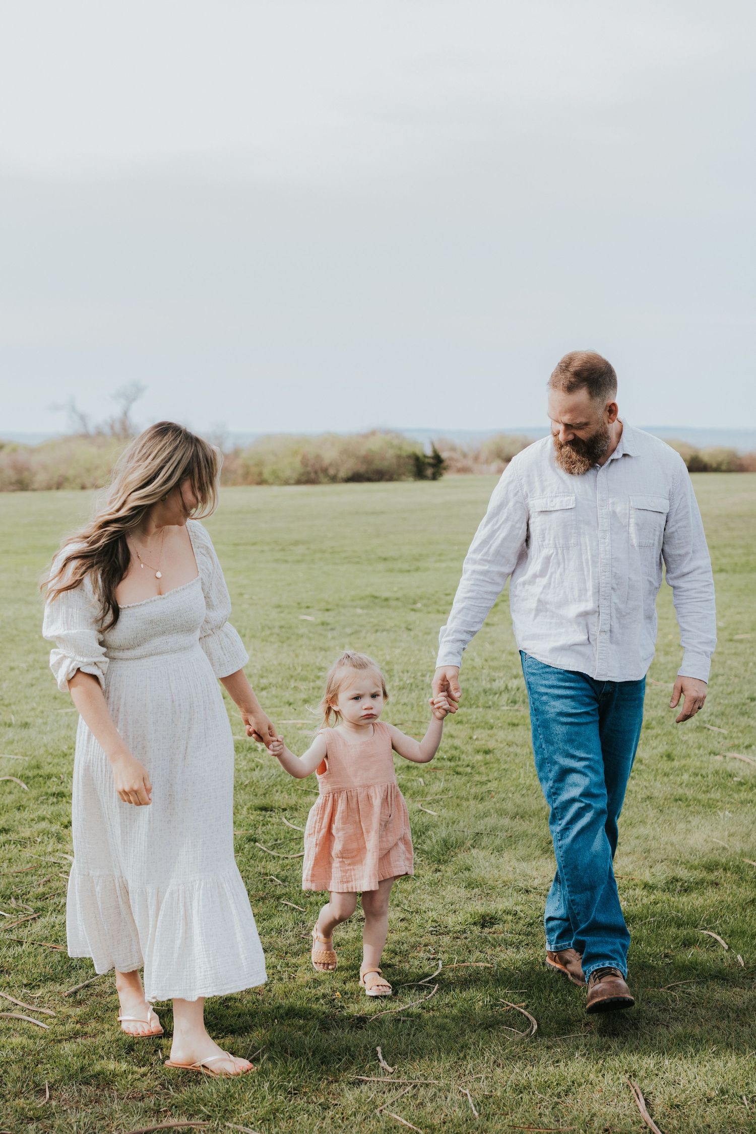 Young family walking on grass, mother in white dress and father in blue jeans holding toddler daughter's hands.