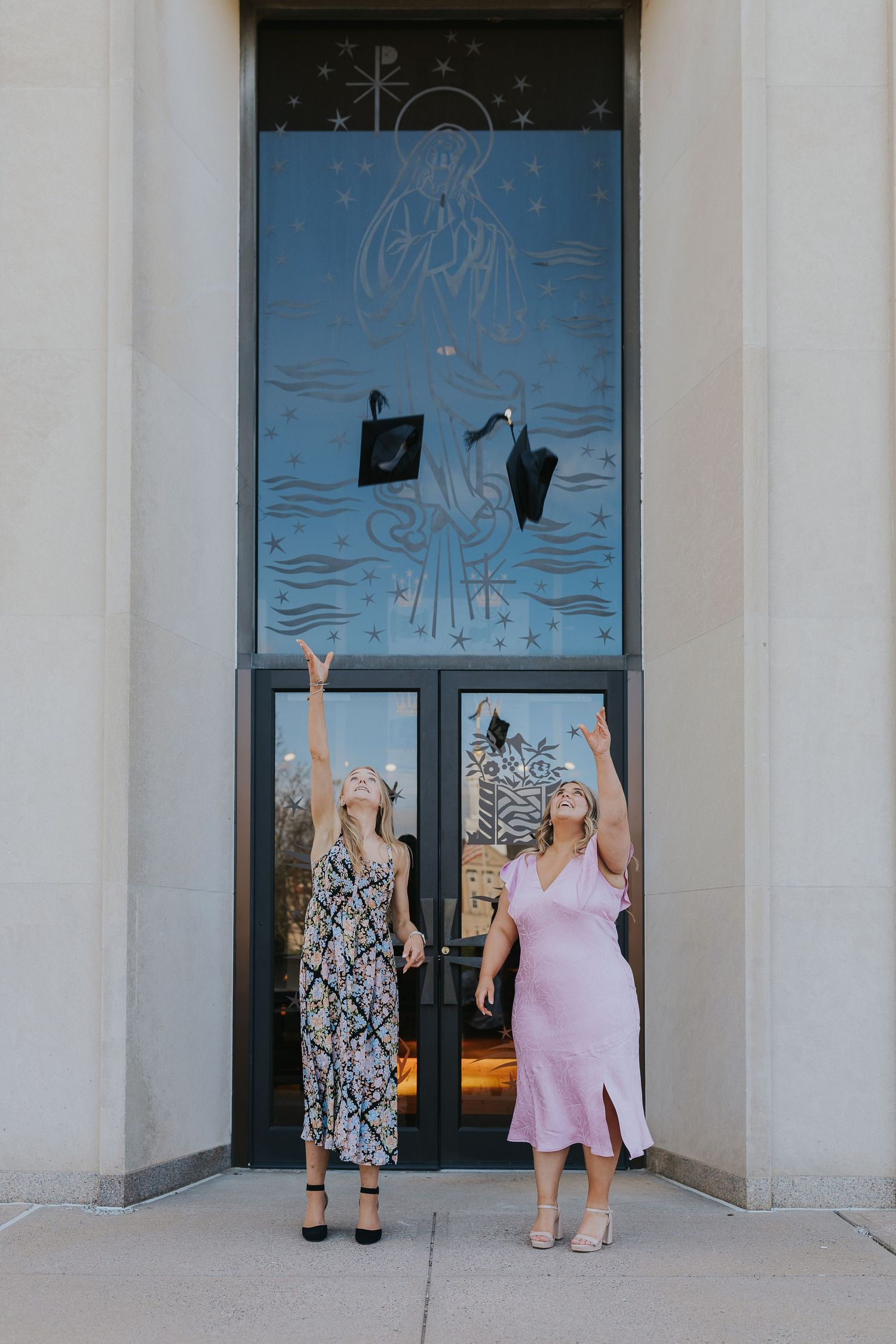 Two graduates tossing caps in air outside decorated building entrance.