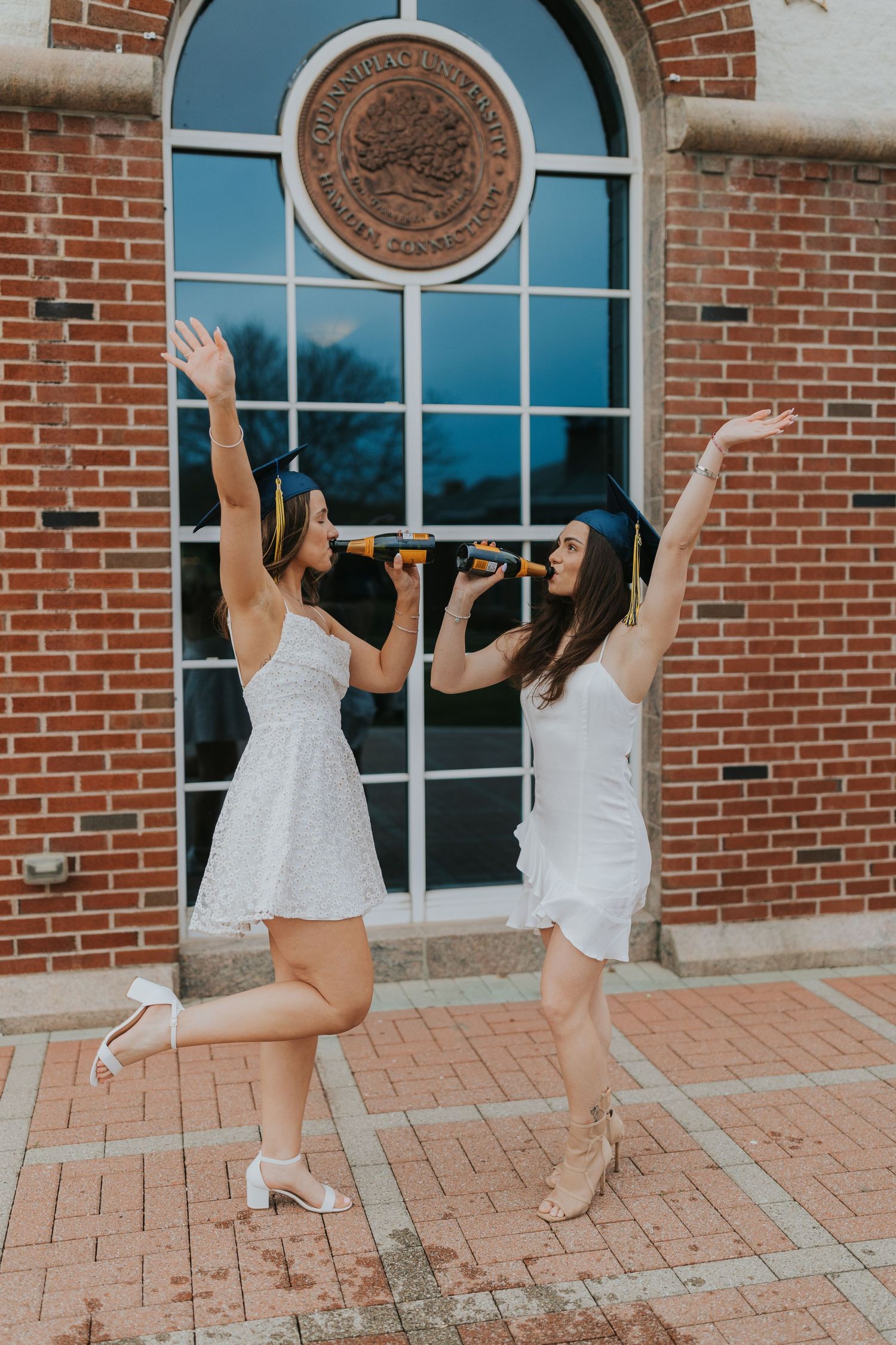 Two graduates in white dresses celebrate joyfully outside Quinnipiac University brick building entrance.