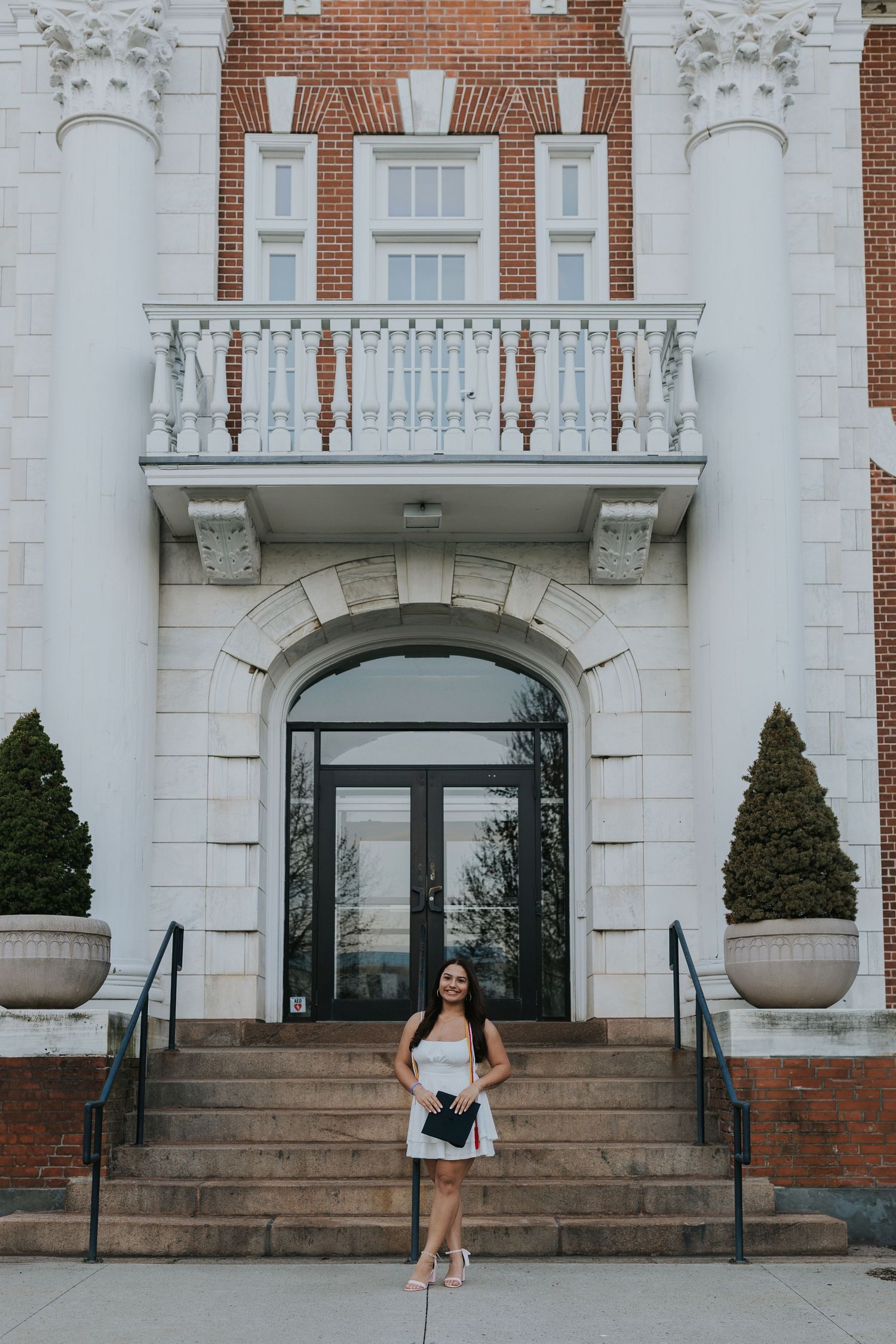 Smiling graduate in white dress stands before elegant brick and white stone campus building.