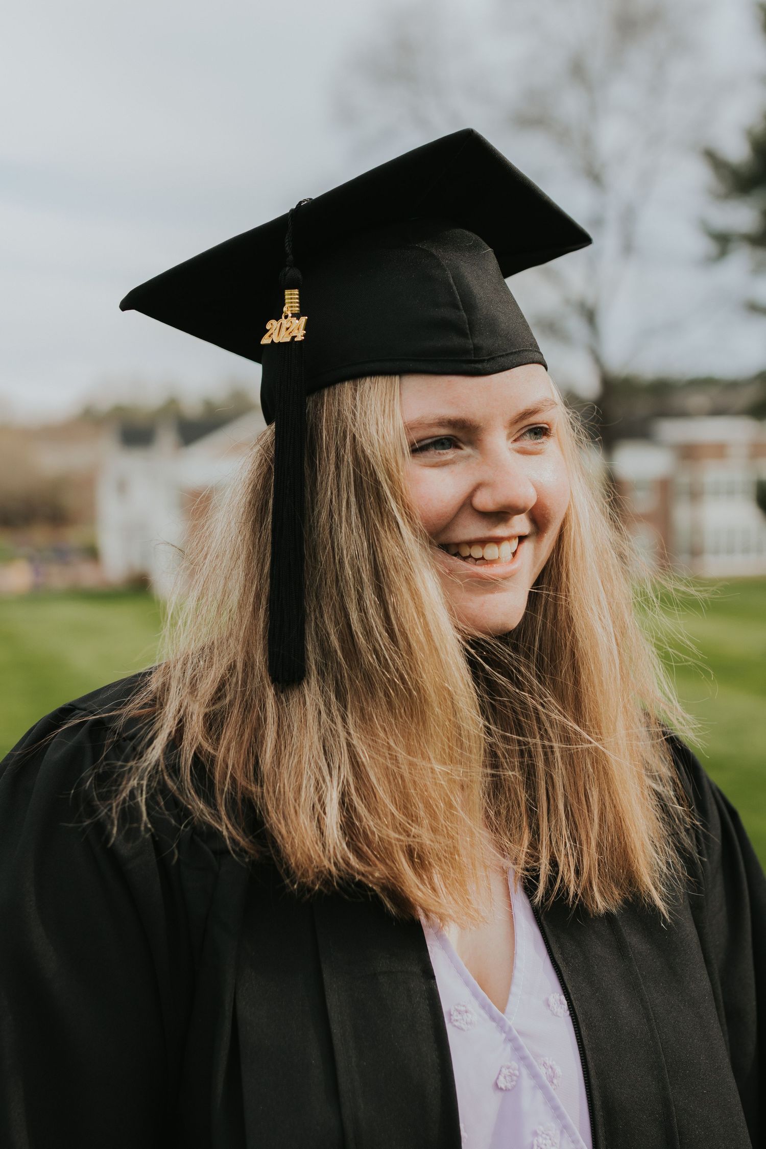 Smiling female graduate in black cap and gown with 2024 tassel outdoors on campus.
