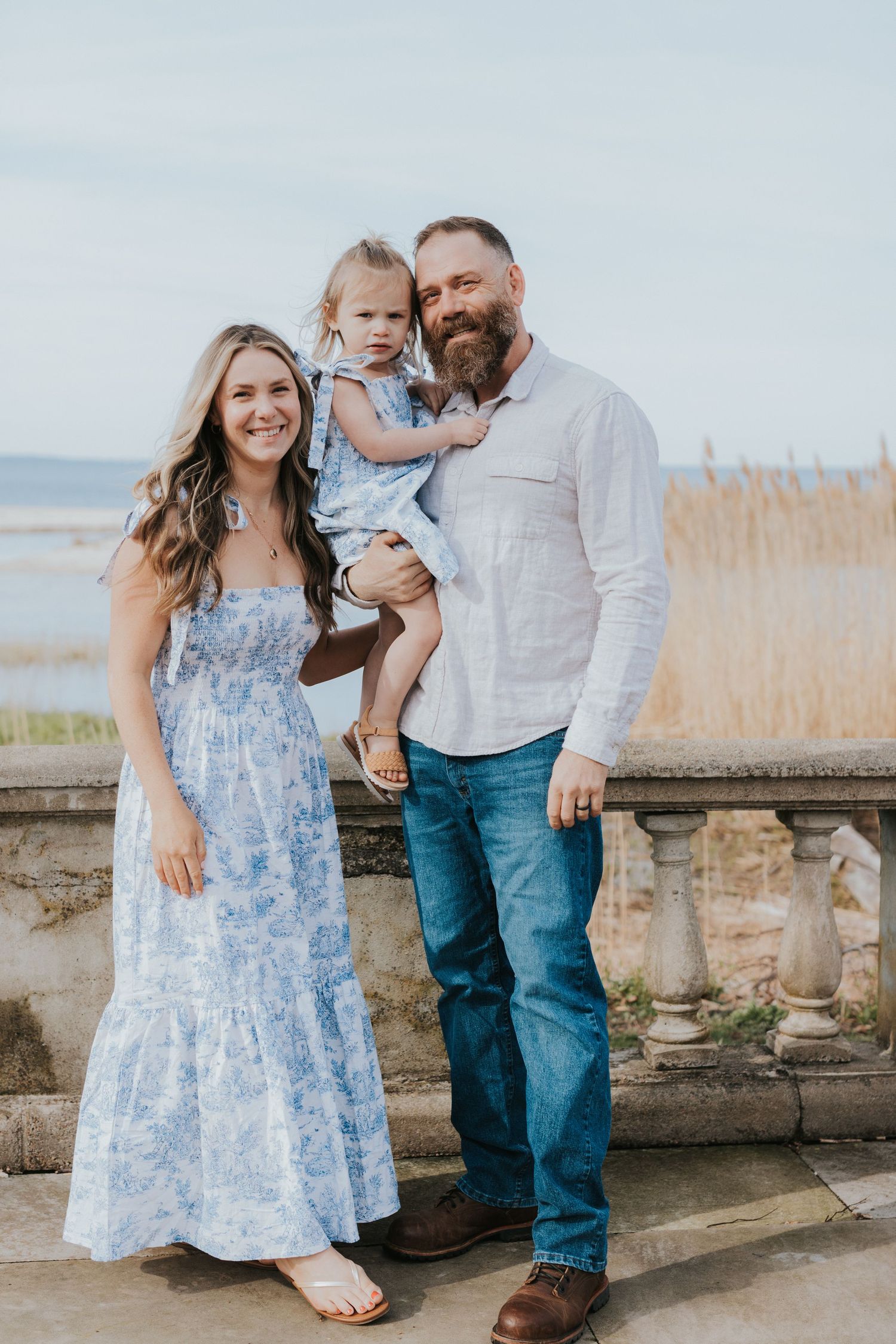 Smiling family portrait with father holding toddler girl and mother in blue floral dress near waterfront.
