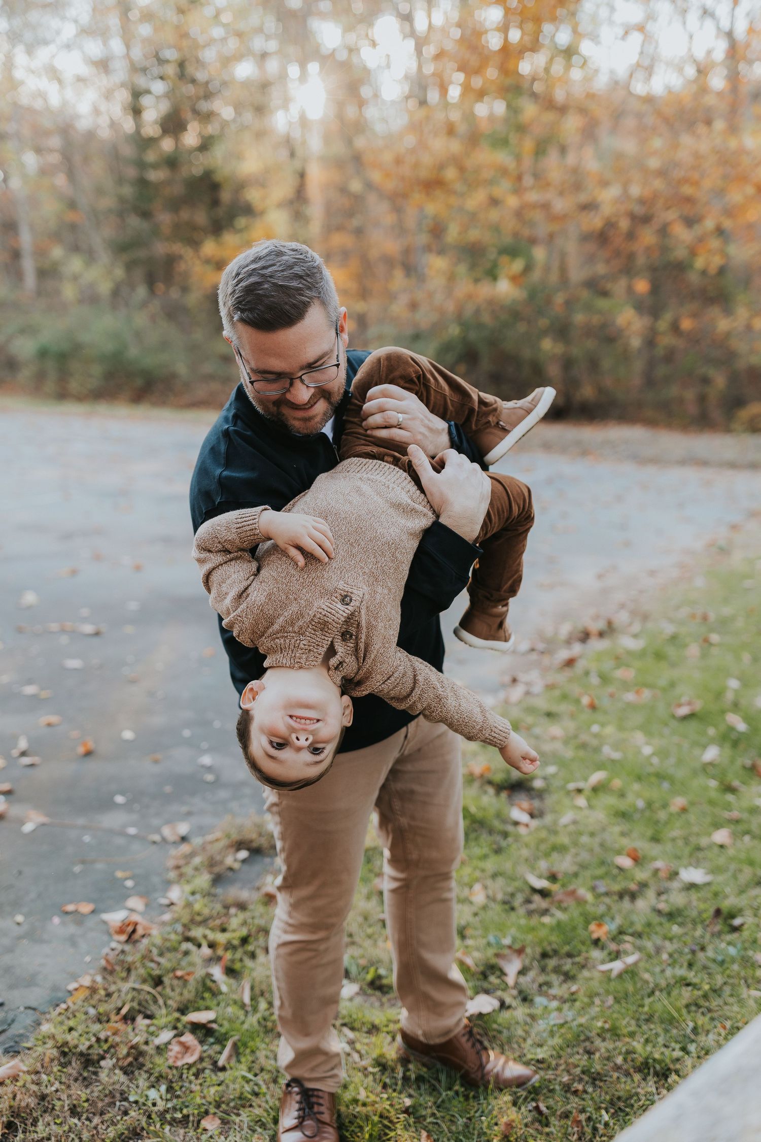 A smiling father playfully holds his young son upside down outdoors during an autumn family photo session.