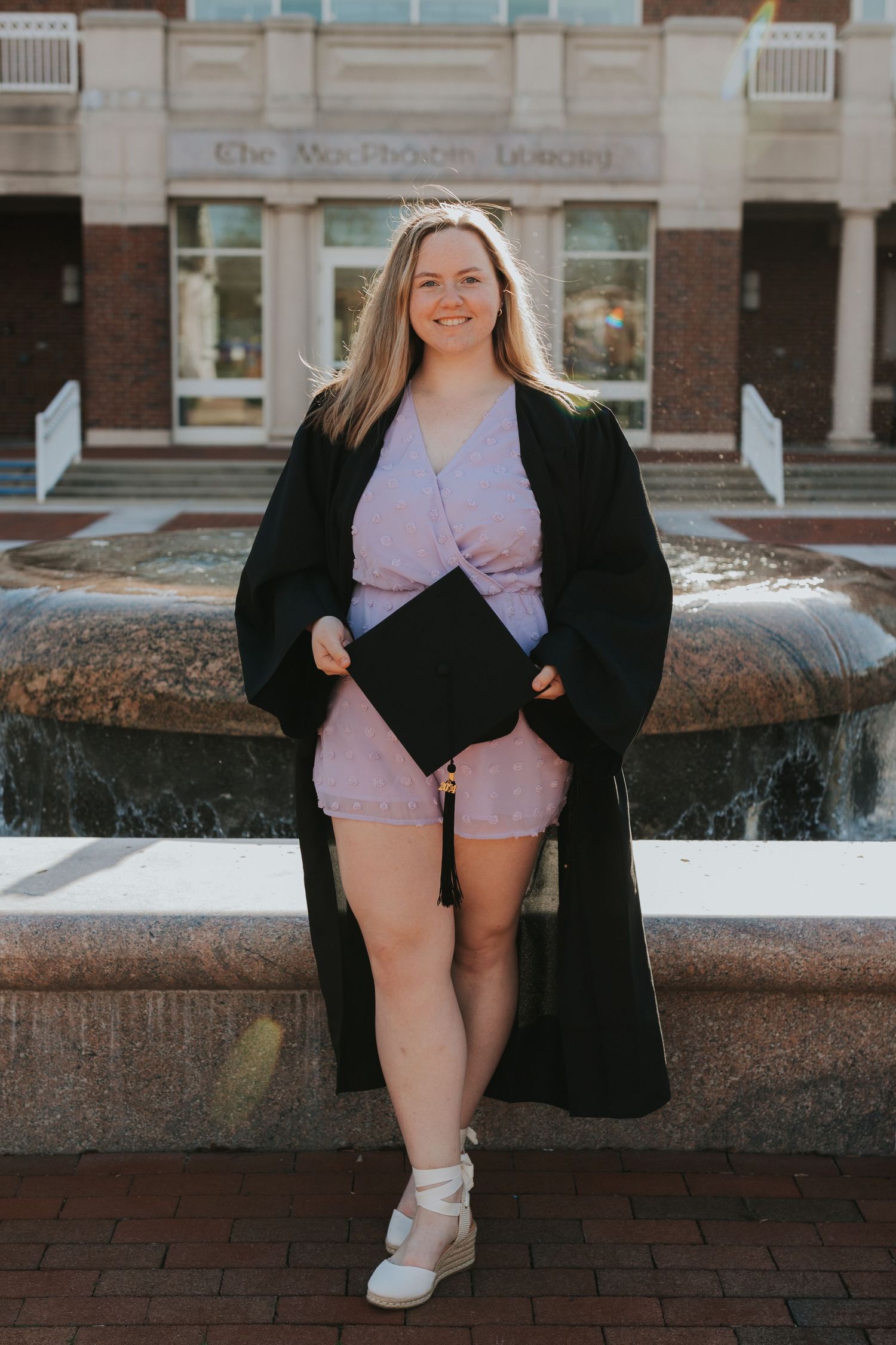 Blonde graduate in black gown holds cap while leaning against fountain outside Washburn Library.