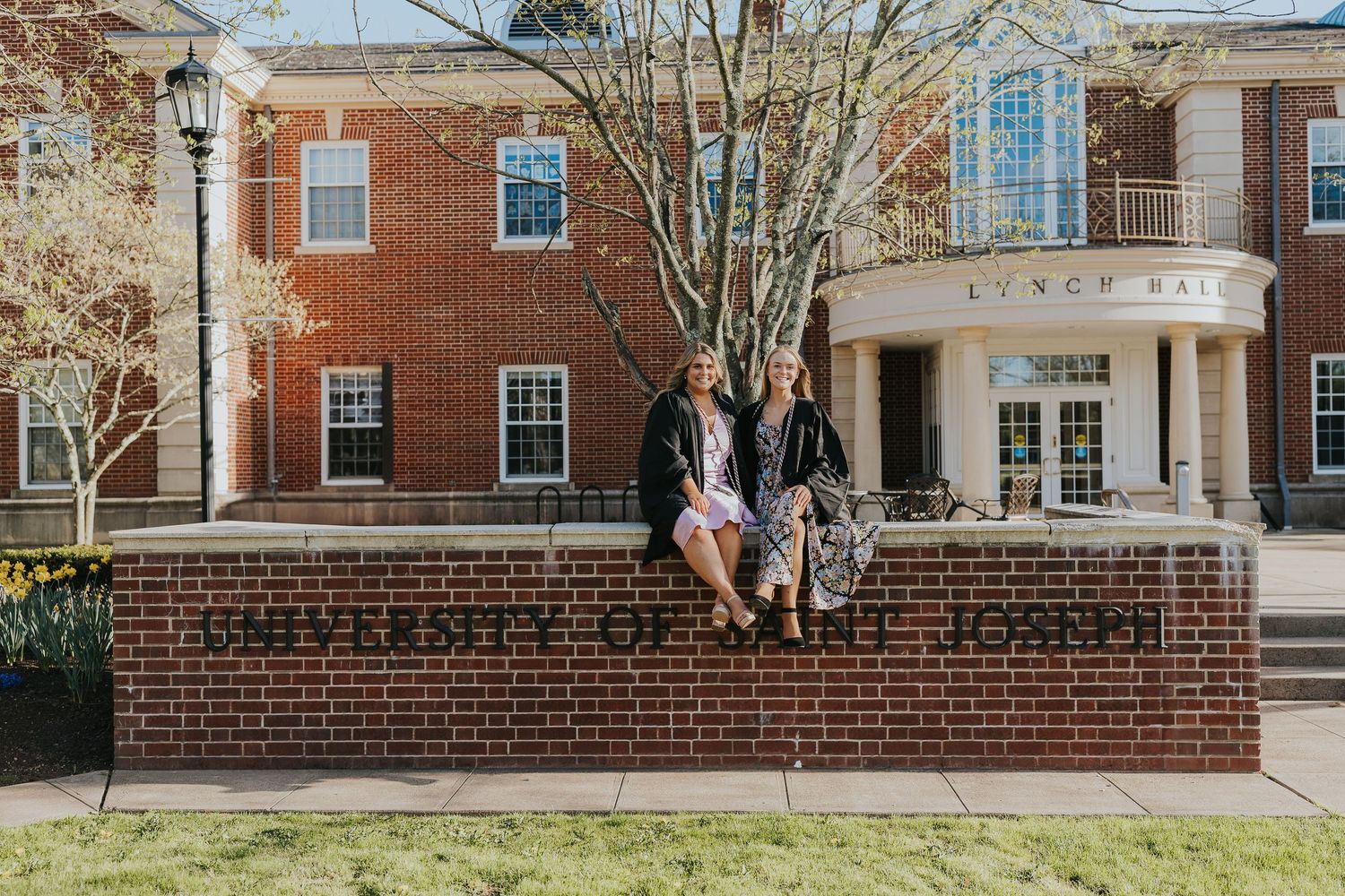 Two graduates in black gowns sitting on University of Saint Joseph sign wall.