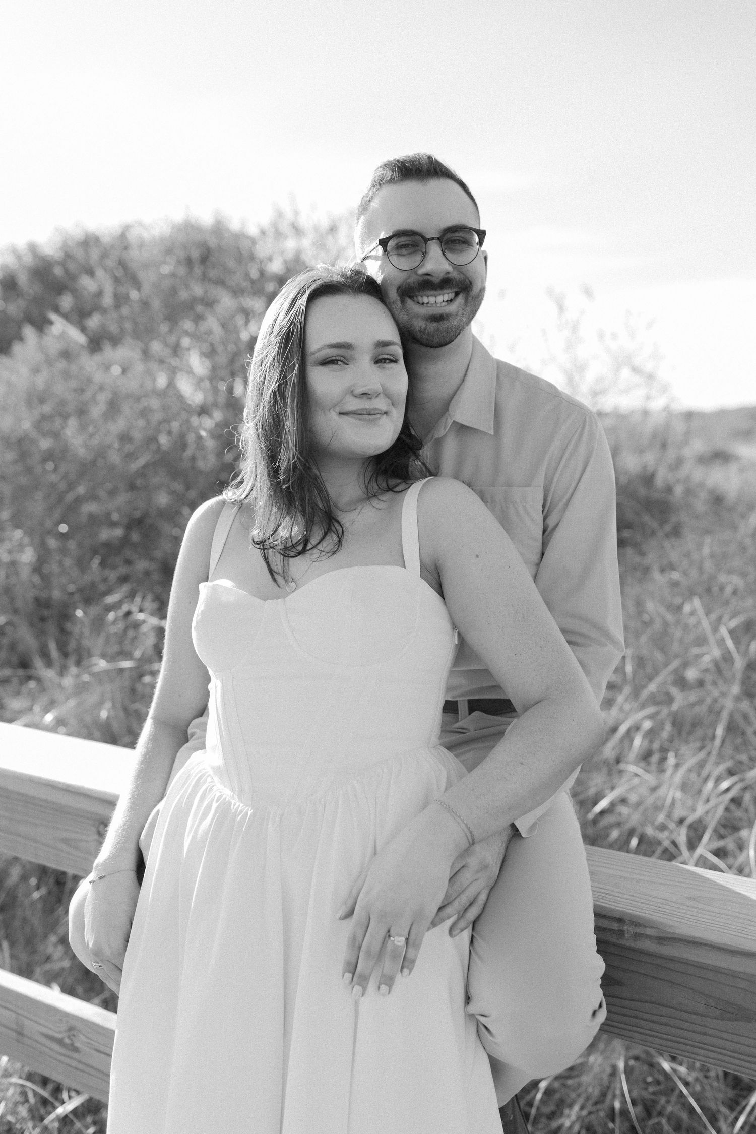 Black and white photo of smiling couple embracing near wooden fence with coastal dunes behind.