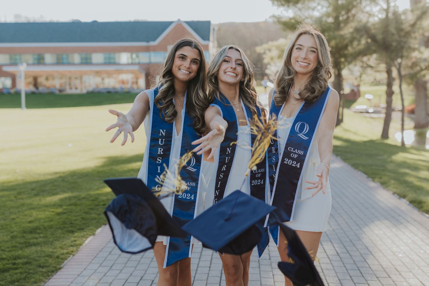 Three smiling college graduates in blue caps and gowns toss their mortarboards outdoors on a sunny campus.