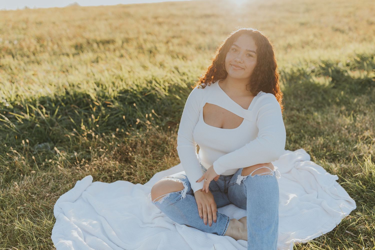 Young woman in white top and jeans sits cross-legged on blanket in sunlit grassy field.