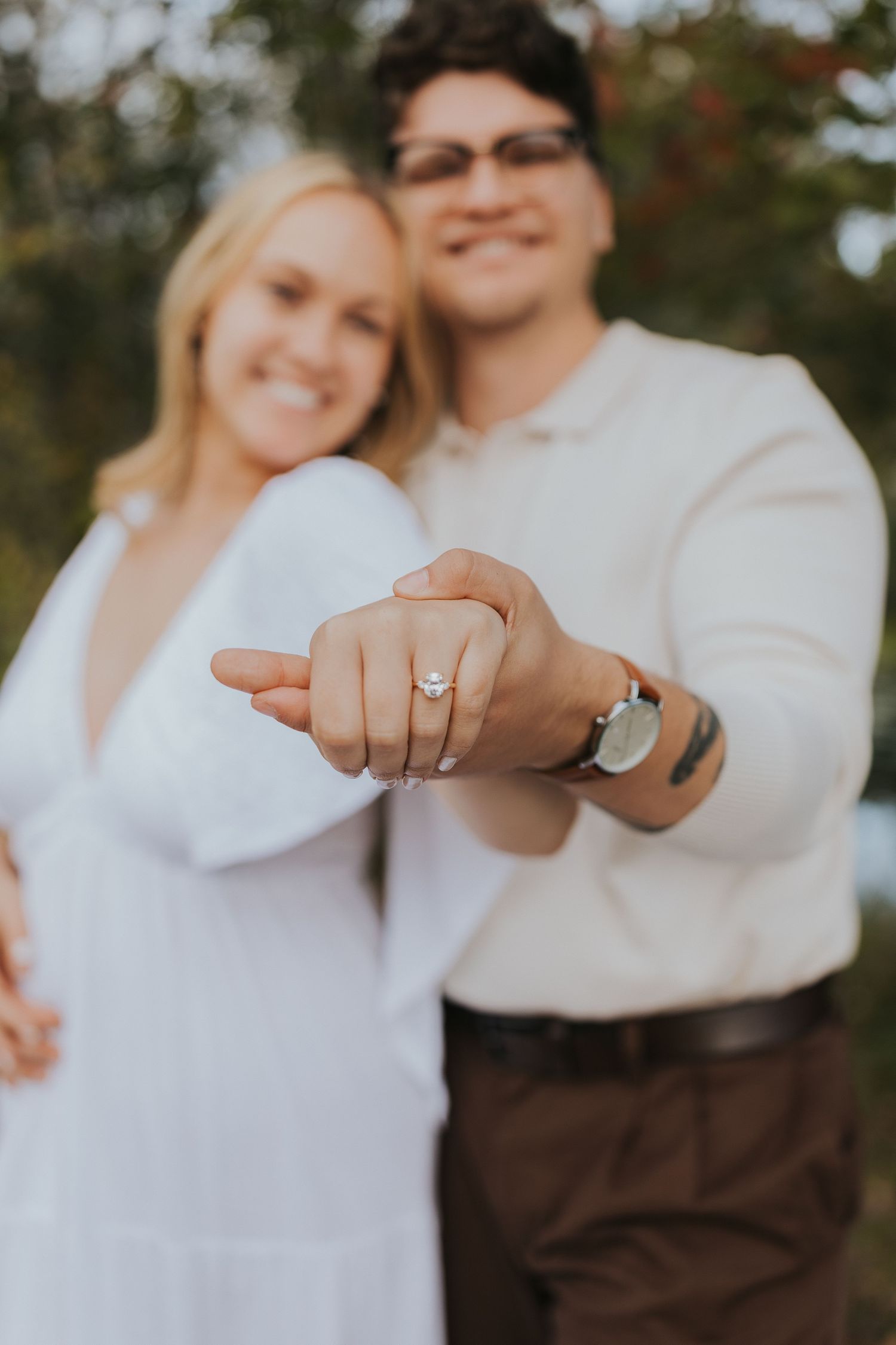Engaged couple smiling and showing off diamond engagement ring in a blurred outdoor autumn setting.