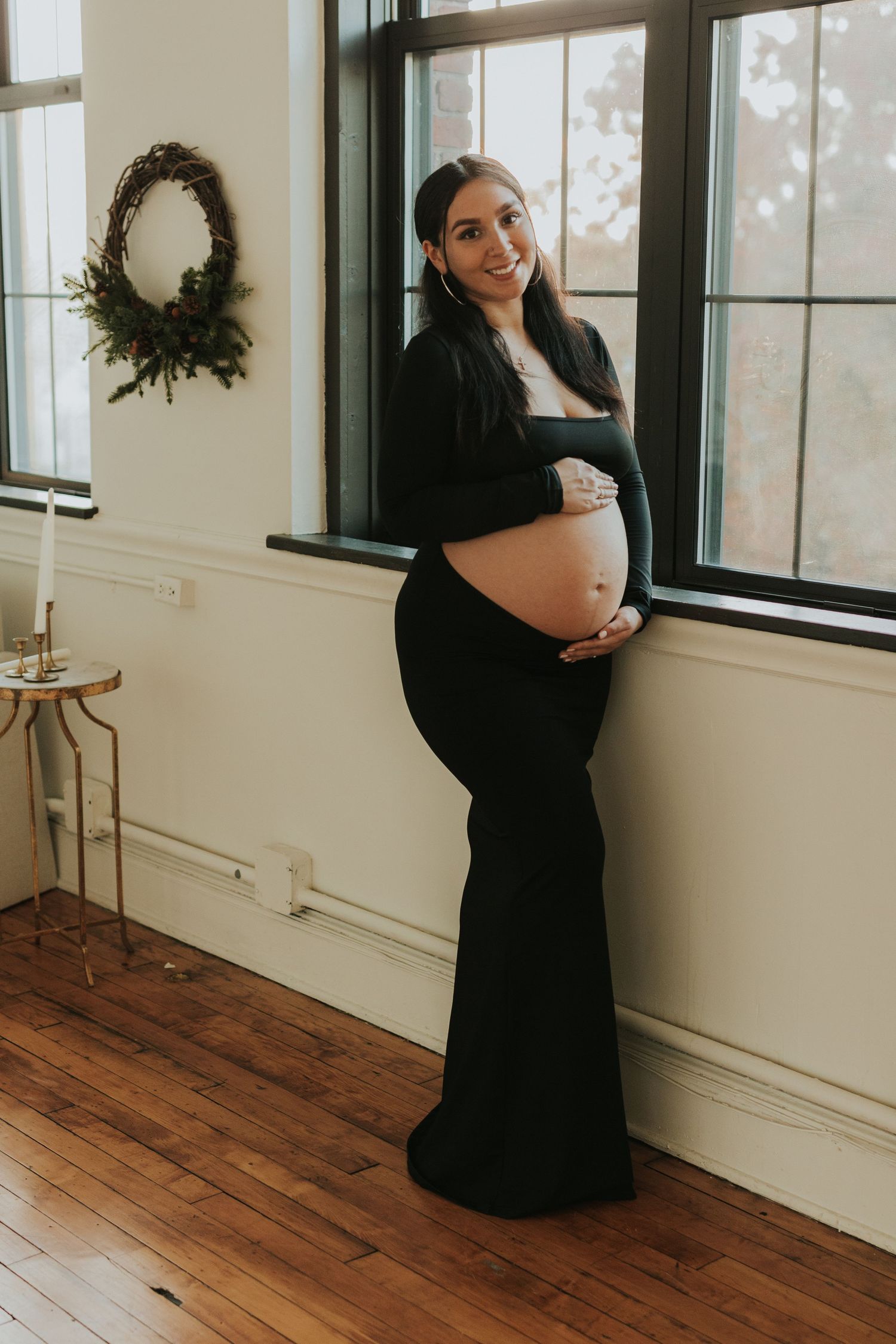 Smiling pregnant woman in black dress cradling baby bump by window with wreath decoration.