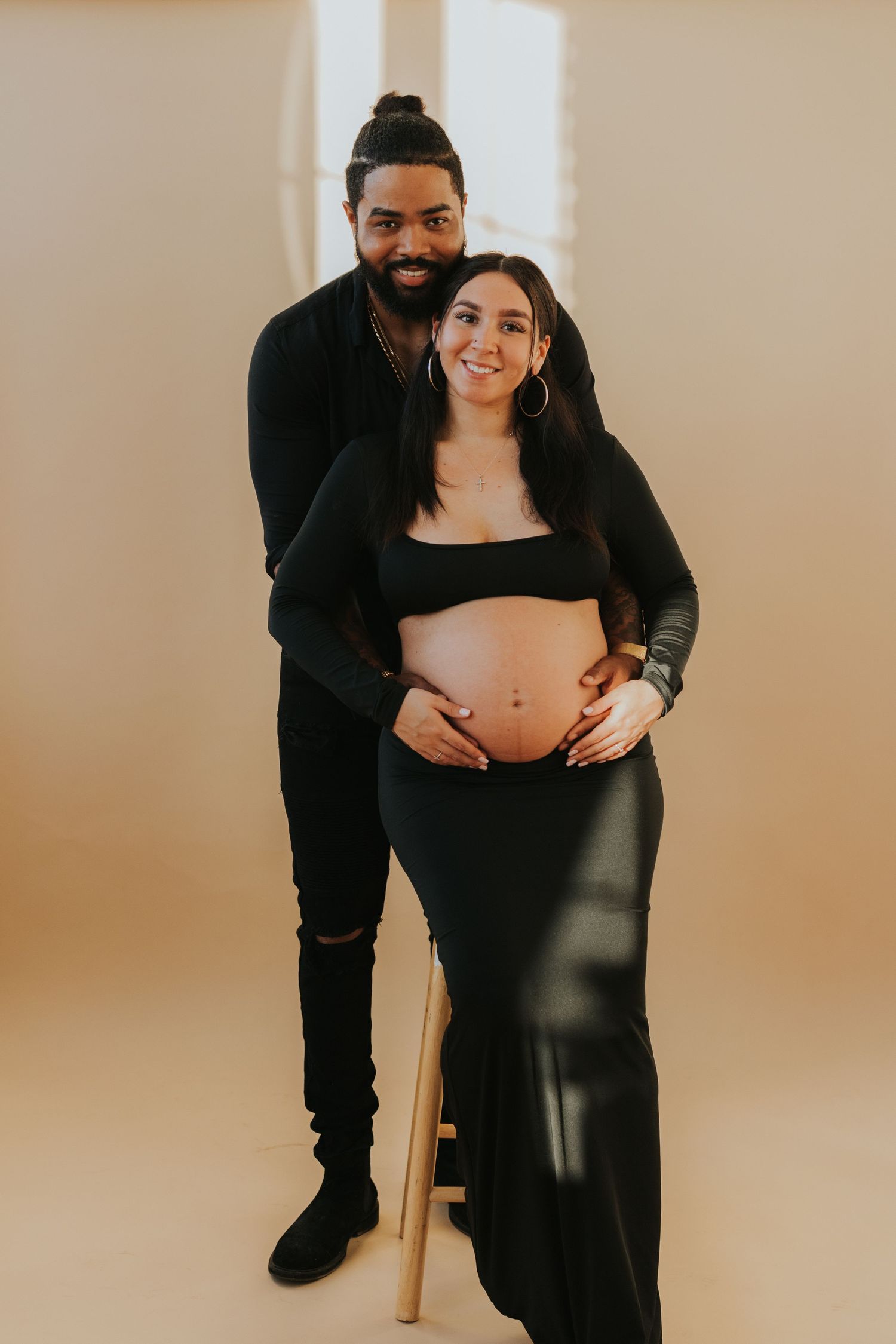 Smiling couple in black outfits posing together with hands on pregnant belly during maternity photoshoot.