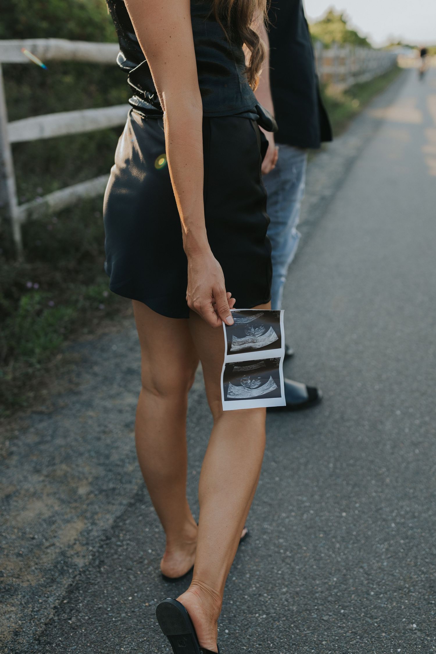 Woman in black dress holding ultrasound photos while walking on a road during a pregnancy announcement photoshoot.