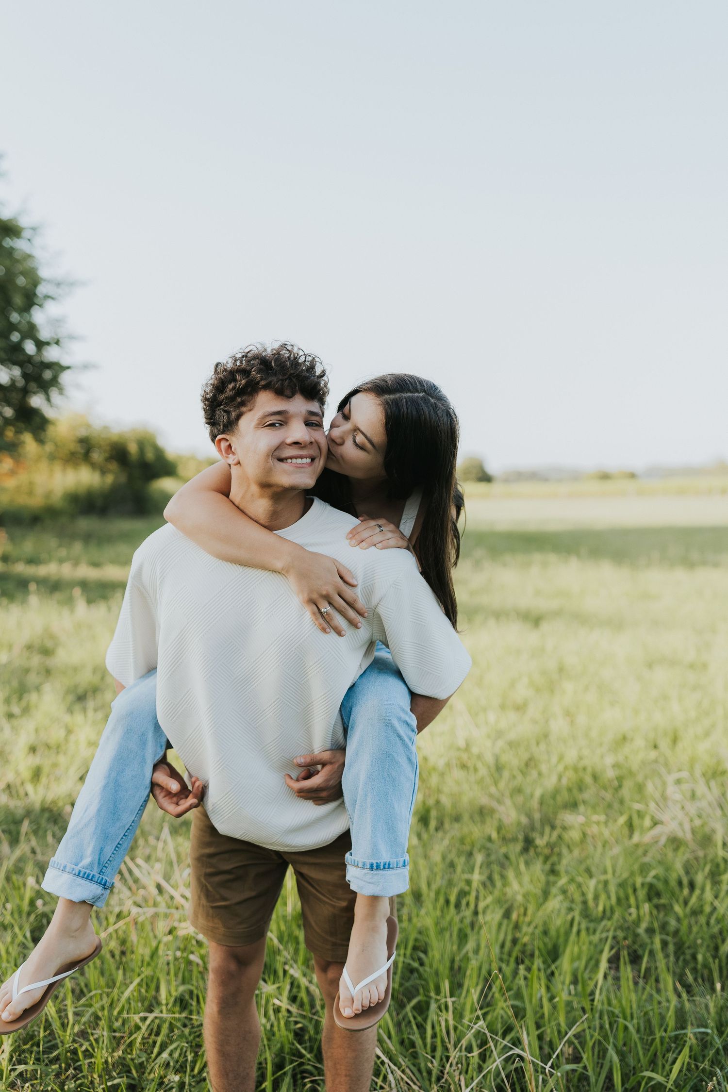 Young woman kissing mans cheek as he gives her a piggyback ride in a green field.