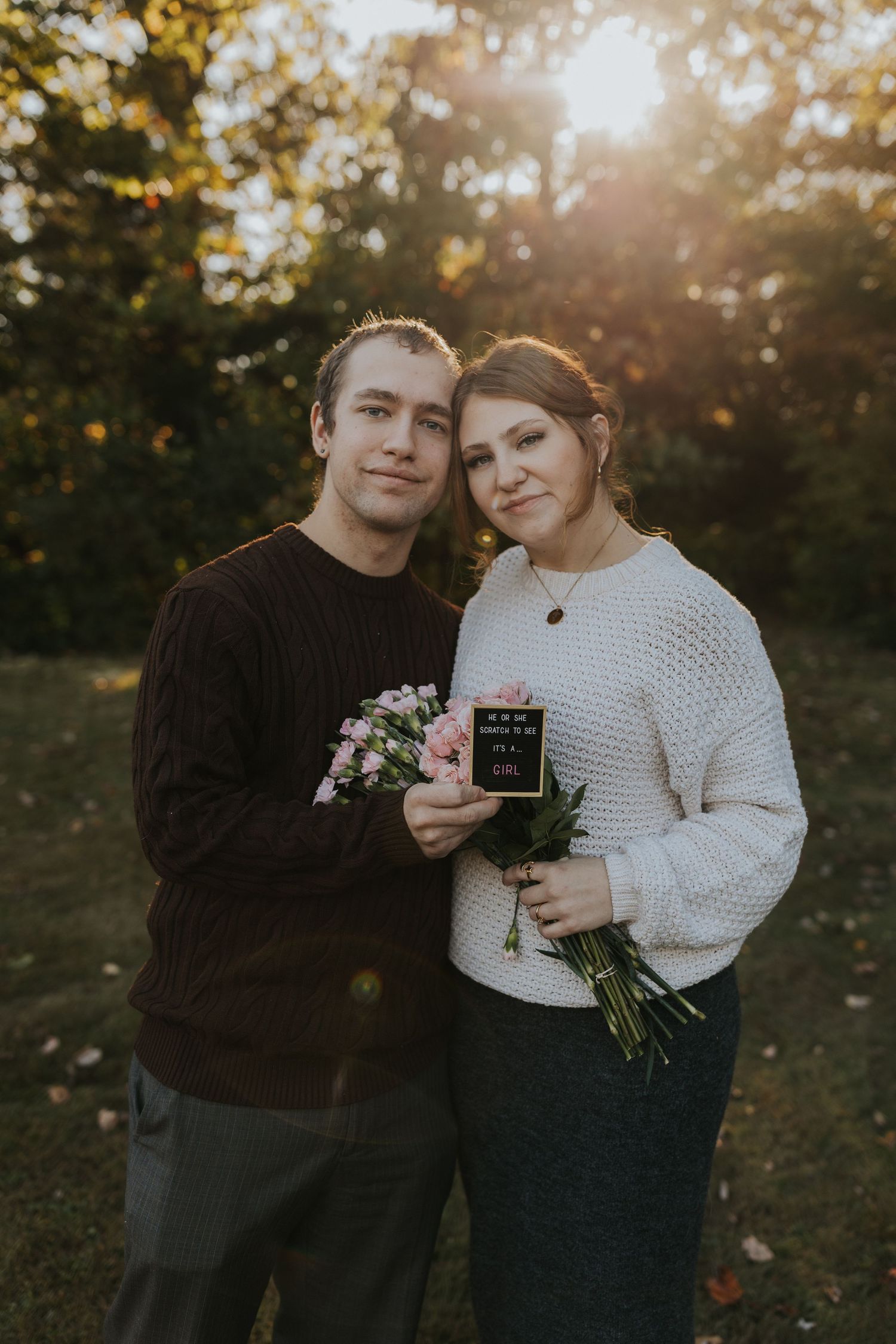 Couple posing with pink bouquet and pregnancy announcement sign during warm sunset outdoor photo session.