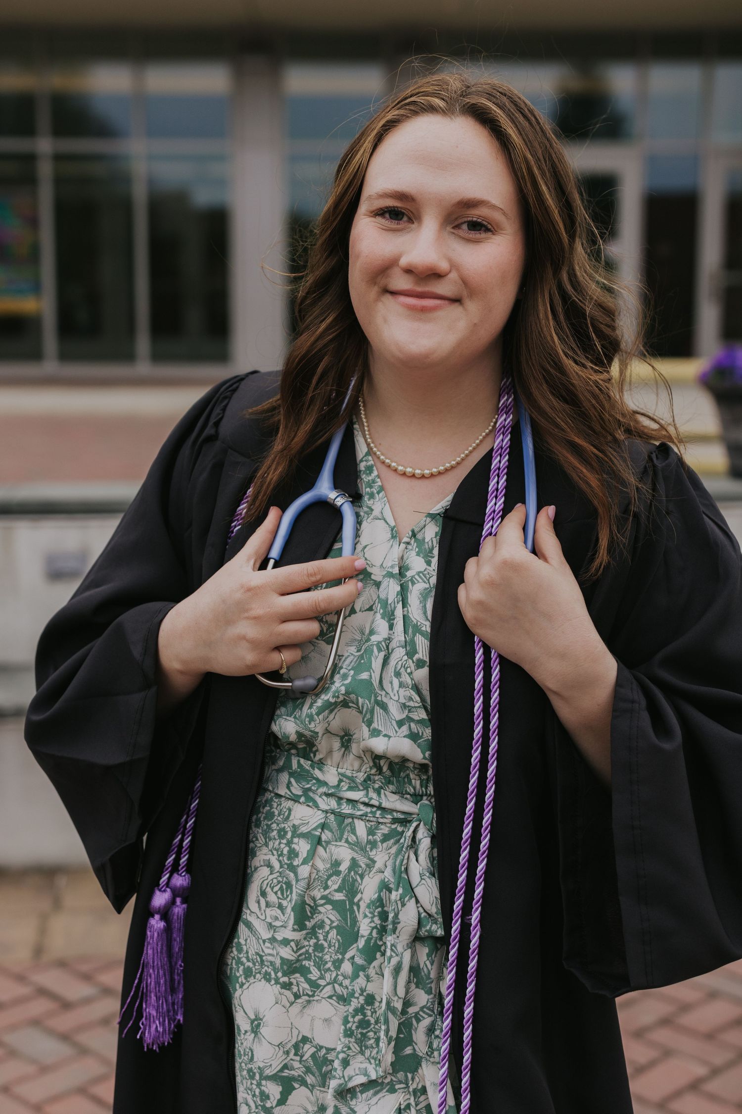 Smiling graduate in black gown with stethoscope and purple honor cord outside library.