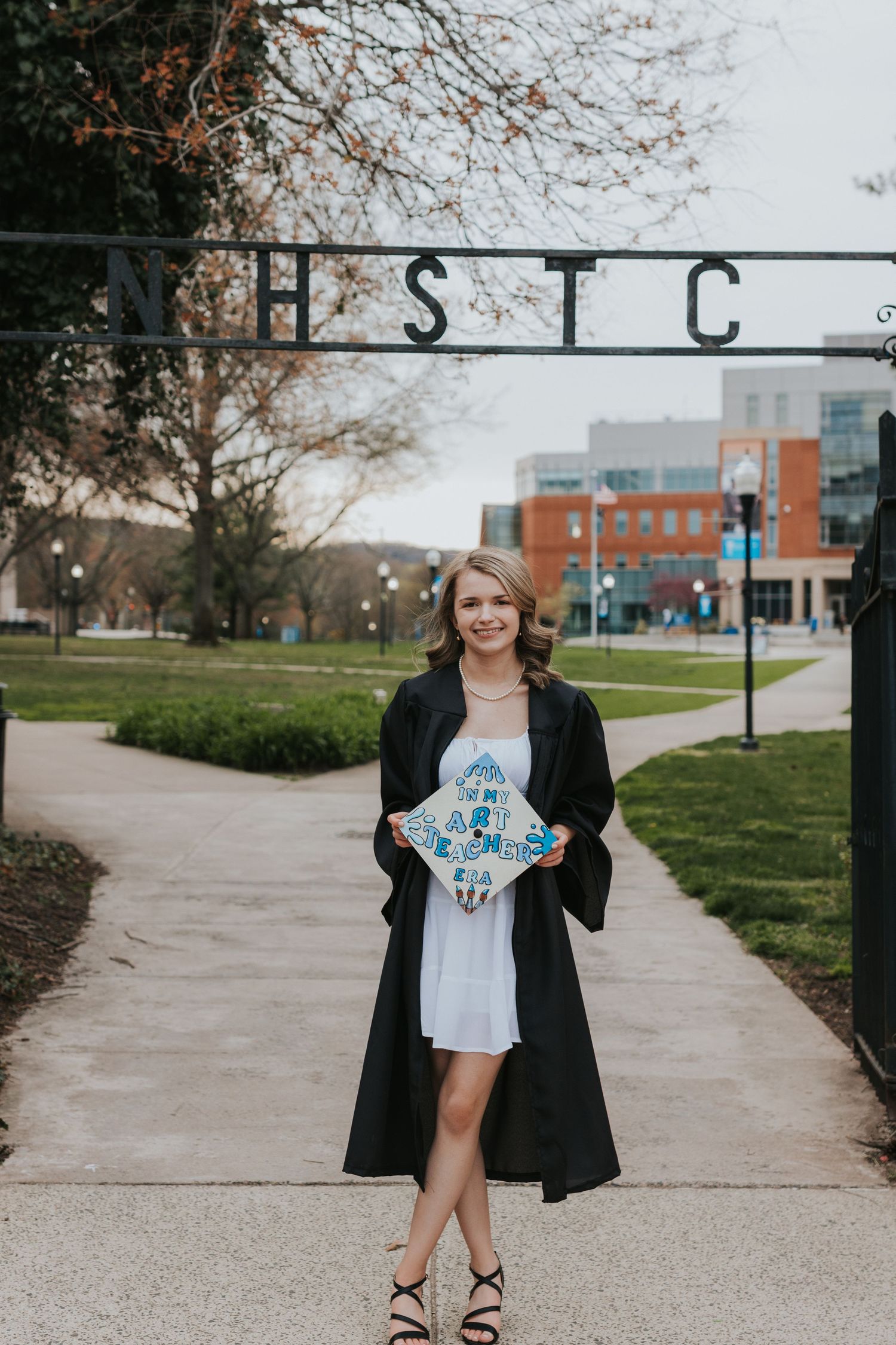 Smiling female graduate holds decorated cap under NHSTC campus gate archway.