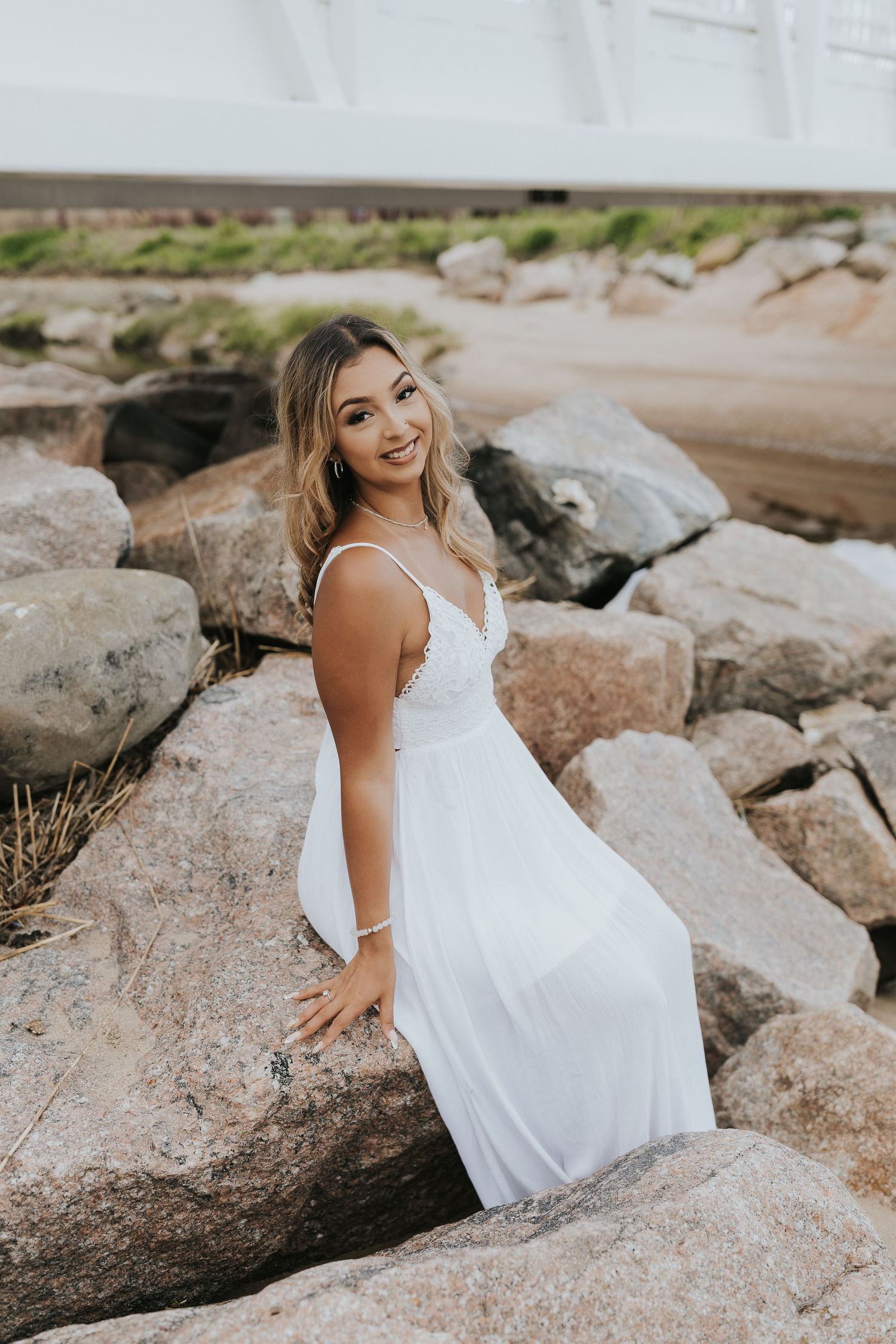 Young woman in white maxi dress sitting on rocky shoreline smiling over shoulder.