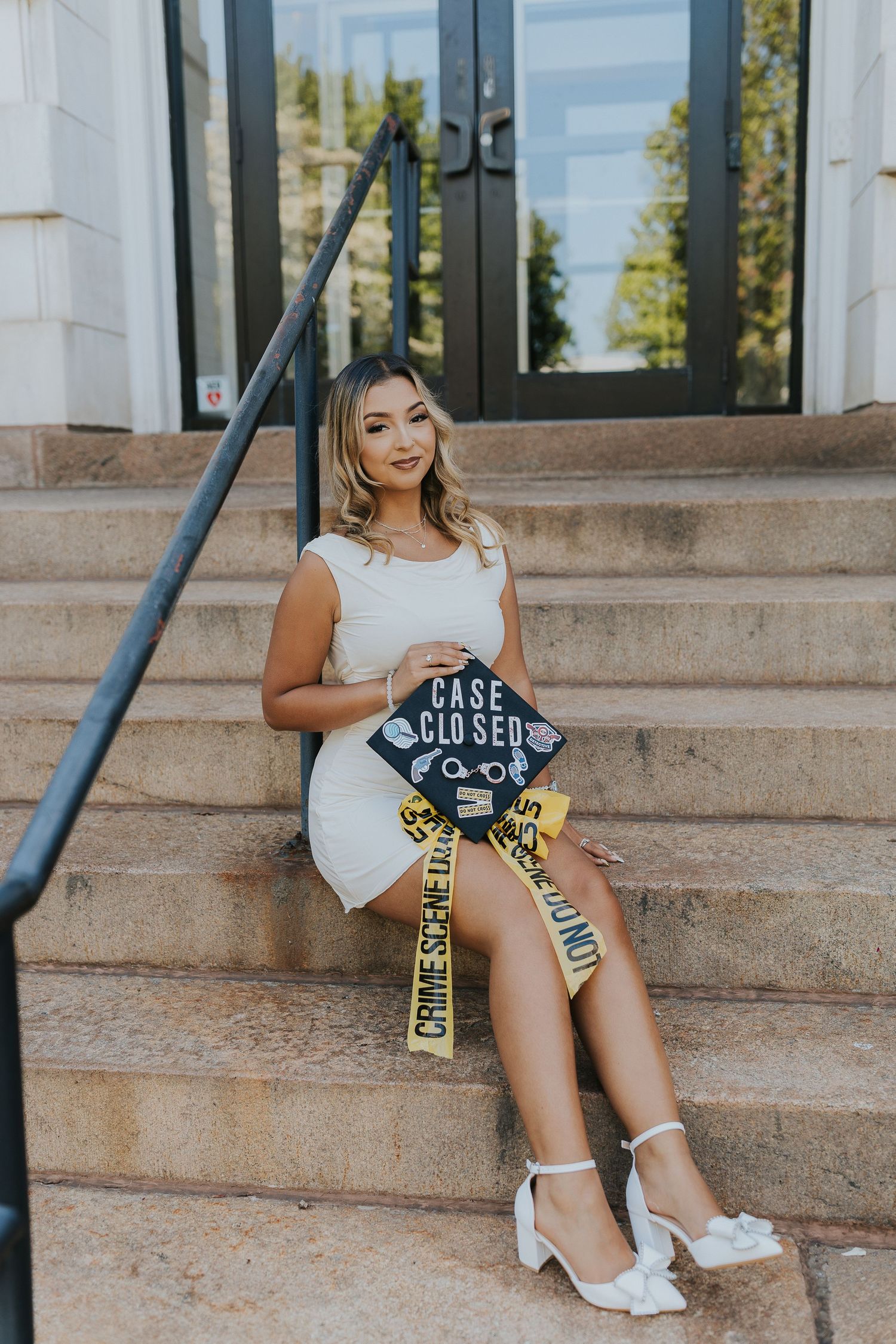 Graduate in white dress sits on stone steps holding a decorated Case Closed forensic science graduation cap with crime scene tape.