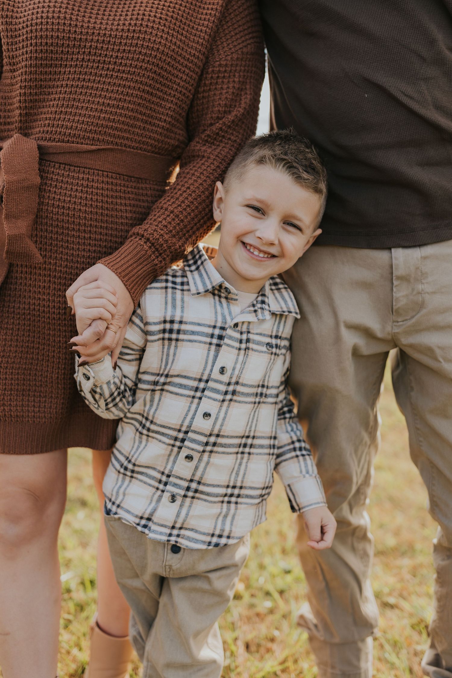 Smiling young boy in plaid shirt holds parents hands while standing outdoors in autumn.