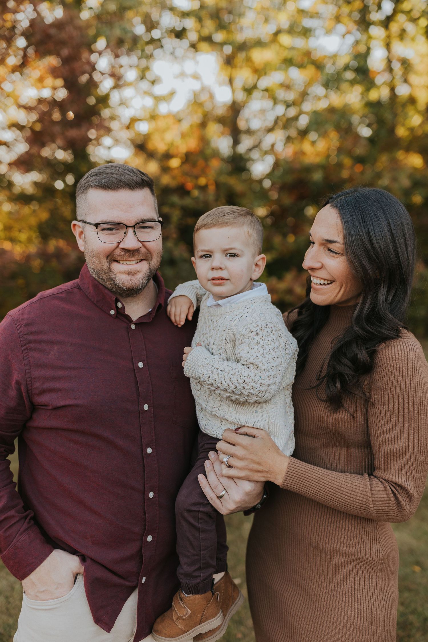Family of three posing outdoors in fall with father in burgundy shirt, mother in brown dress, toddler.