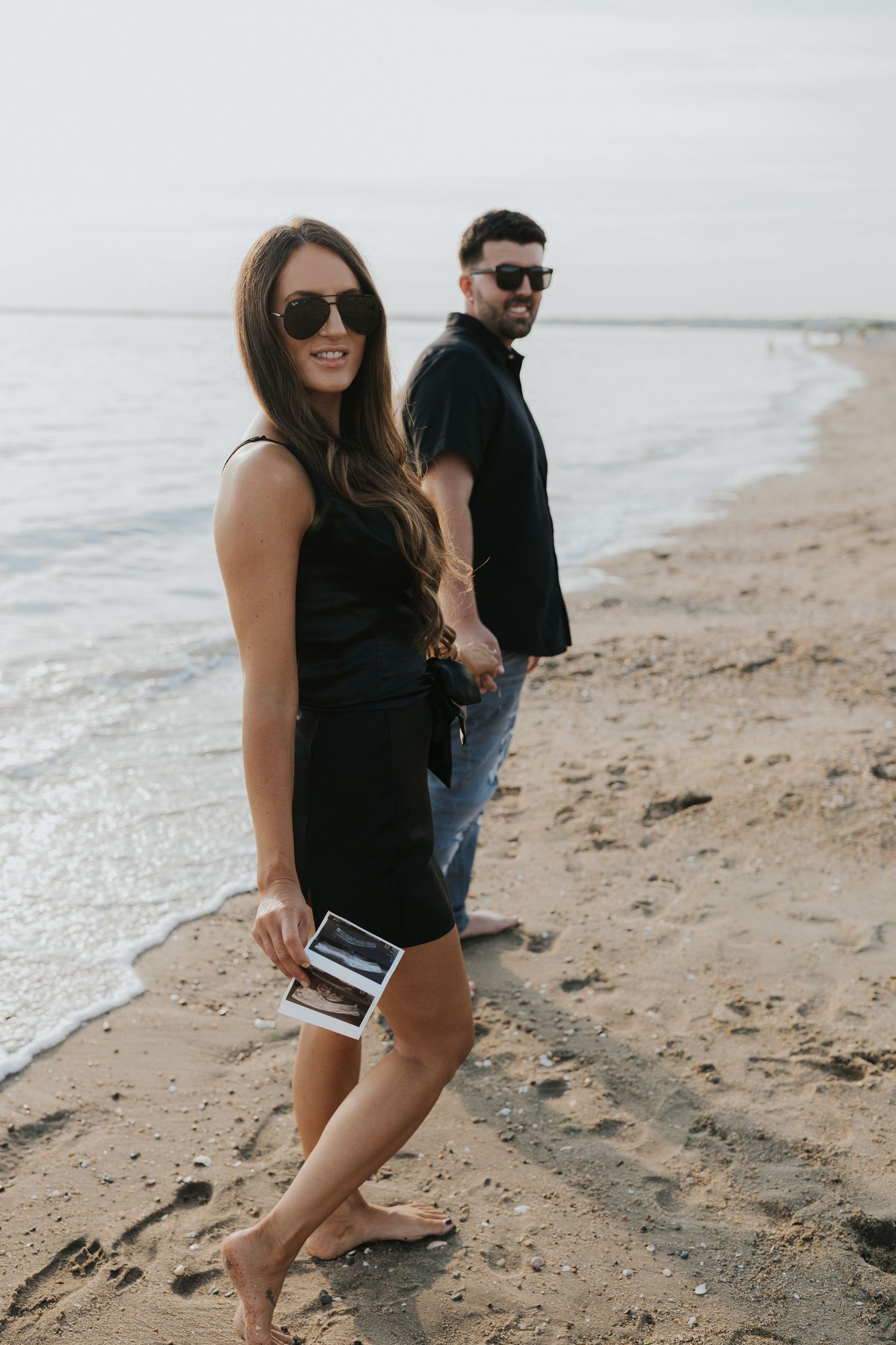 Smiling couple in black outfits holding ultrasound photos while walking barefoot on a sandy beach.