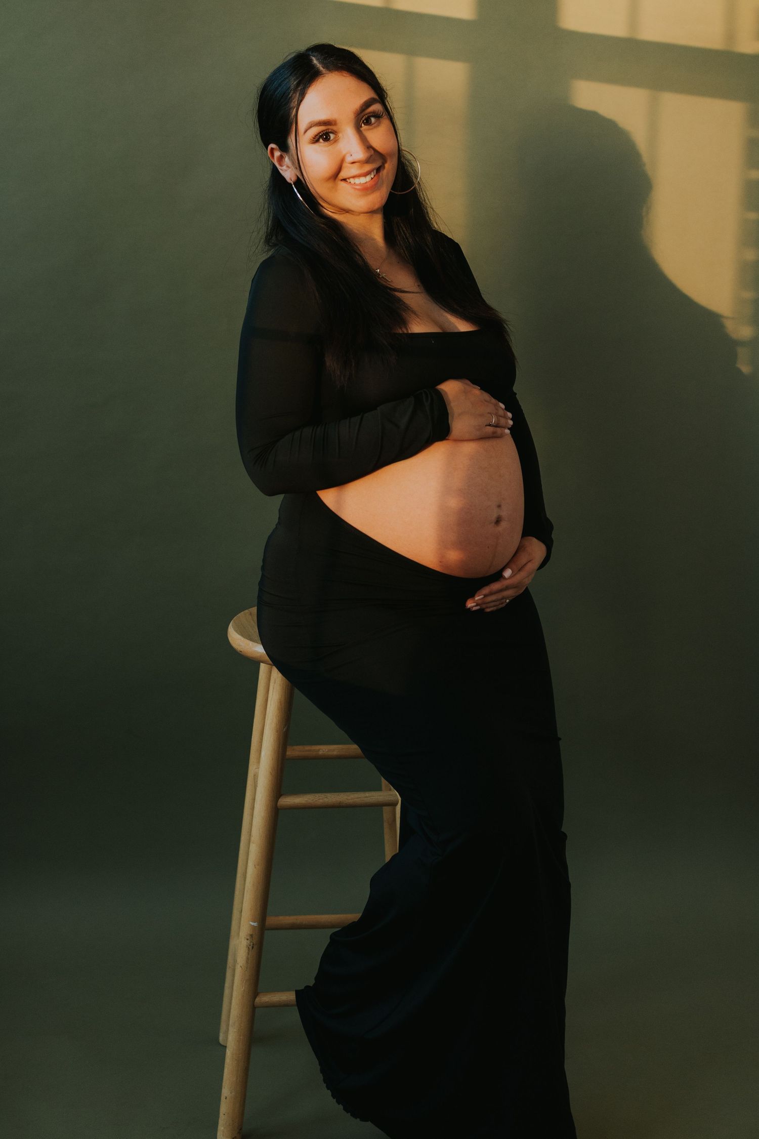 Smiling pregnant woman in black crop top and skirt cradling her baby bump on a stool.
