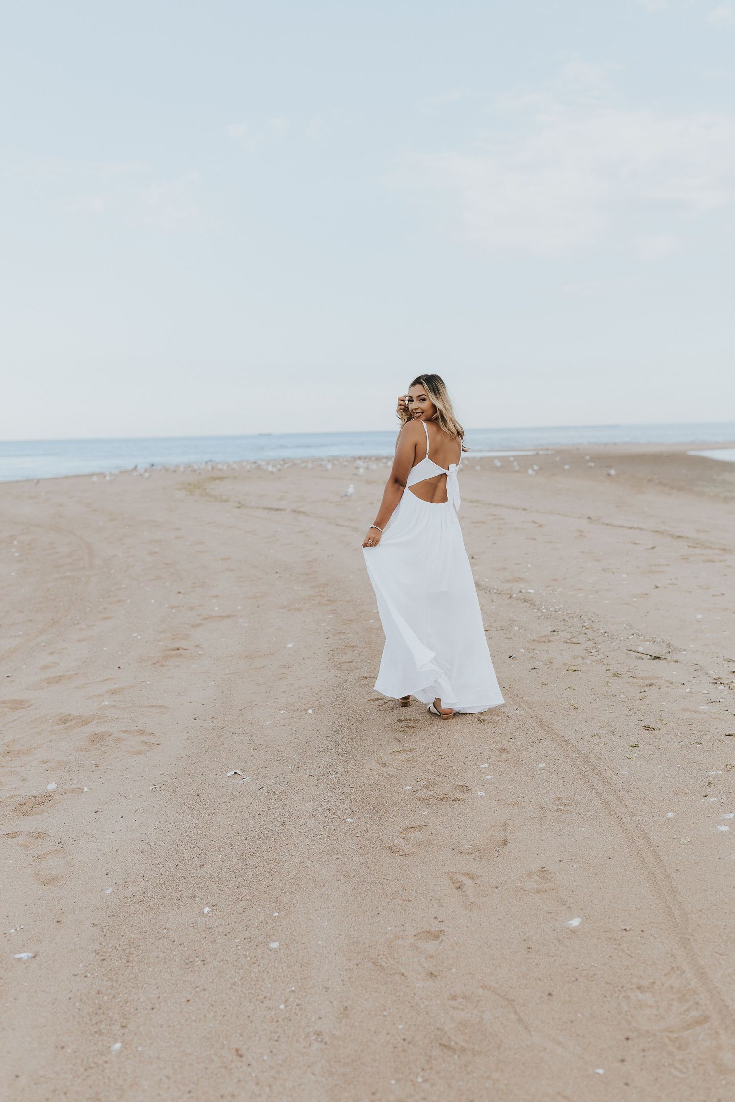 Woman in flowing white dress walking on sandy beach toward ocean.