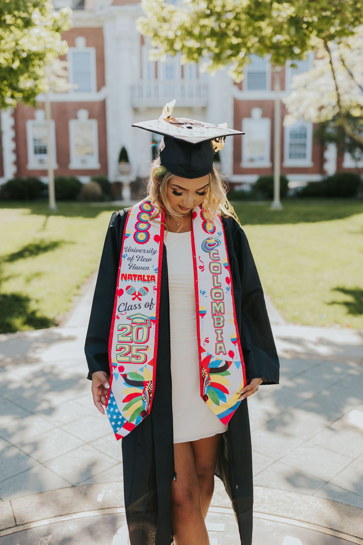 Graduate in black gown and colorful Colombia University of New Haven Class of 2025 stole poses on sunny campus.