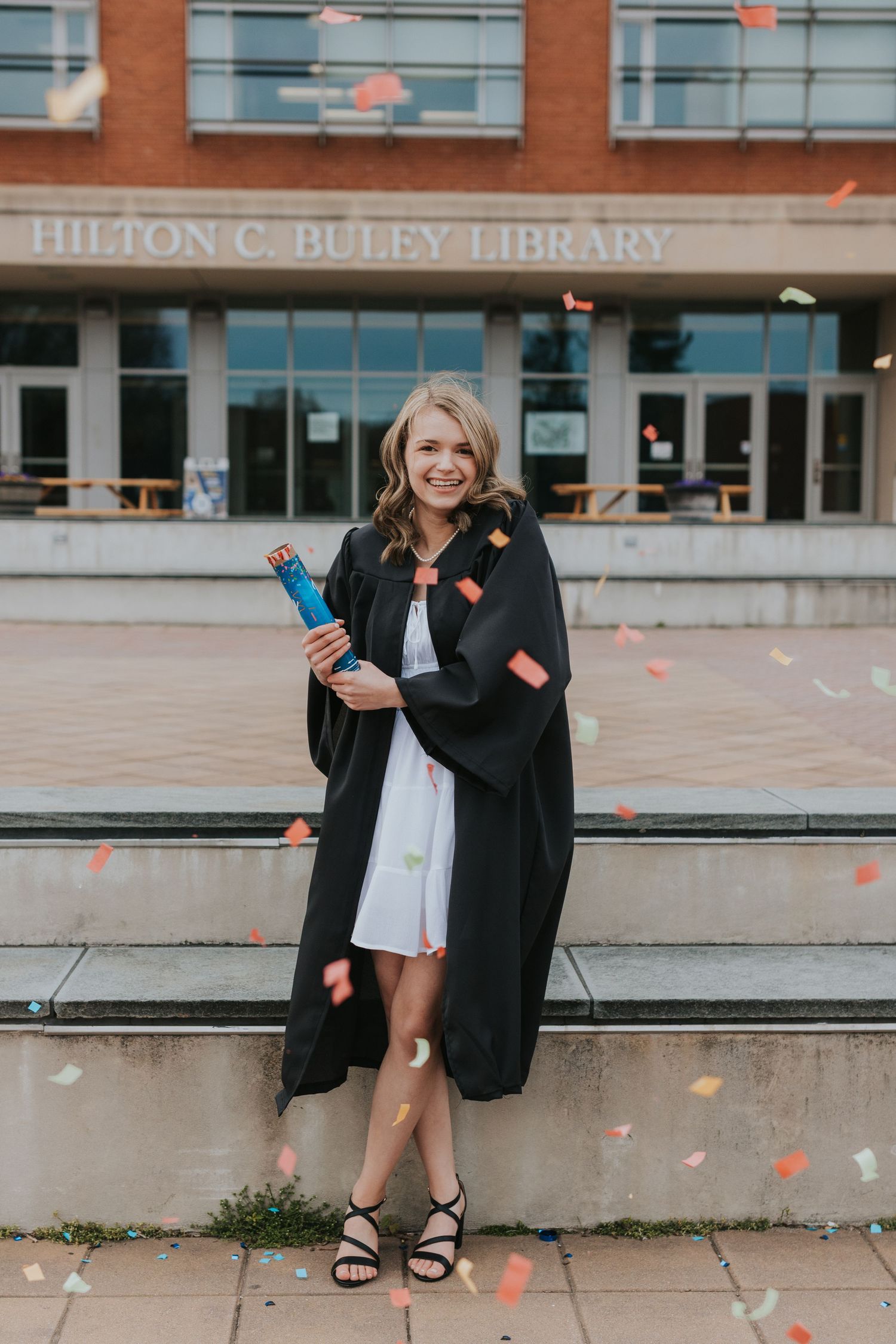 Graduate celebrates with confetti flying outside Hilton C. Buley Library holding blue bottle.