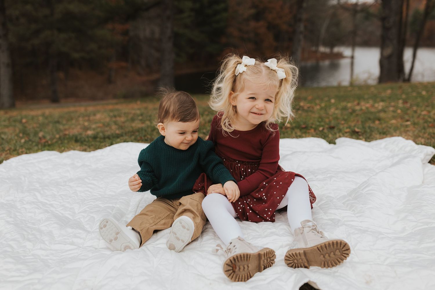 Two young siblings sitting together on a white blanket in an autumn outdoor setting.