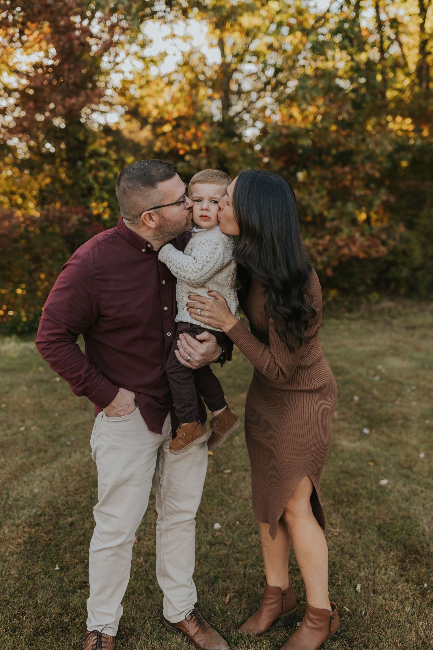 Parents kissing toddler son on cheeks during fall family photo session in autumn park setting.