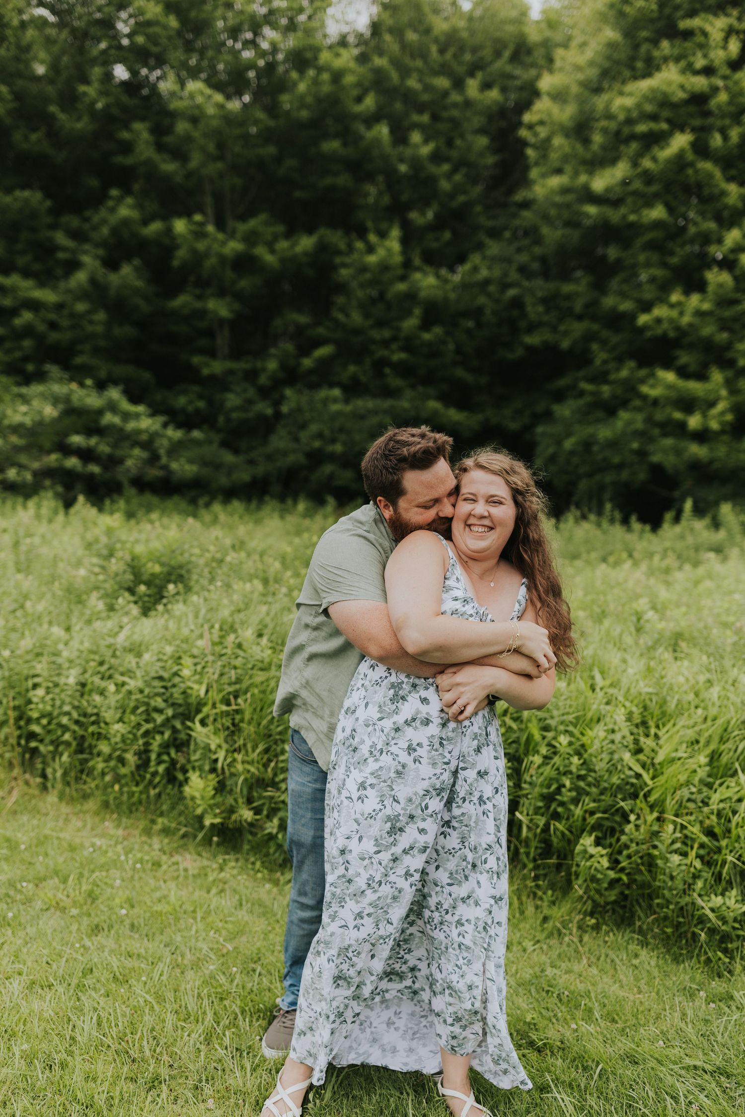 Man hugging woman from behind as she laughs joyfully, standing in a lush green field together.