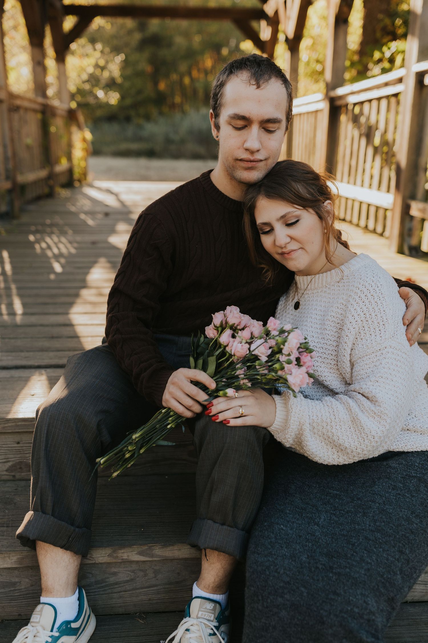 Couple sitting on wooden bridge steps, woman holding pink tulips, man in black sweater embracing her tenderly.