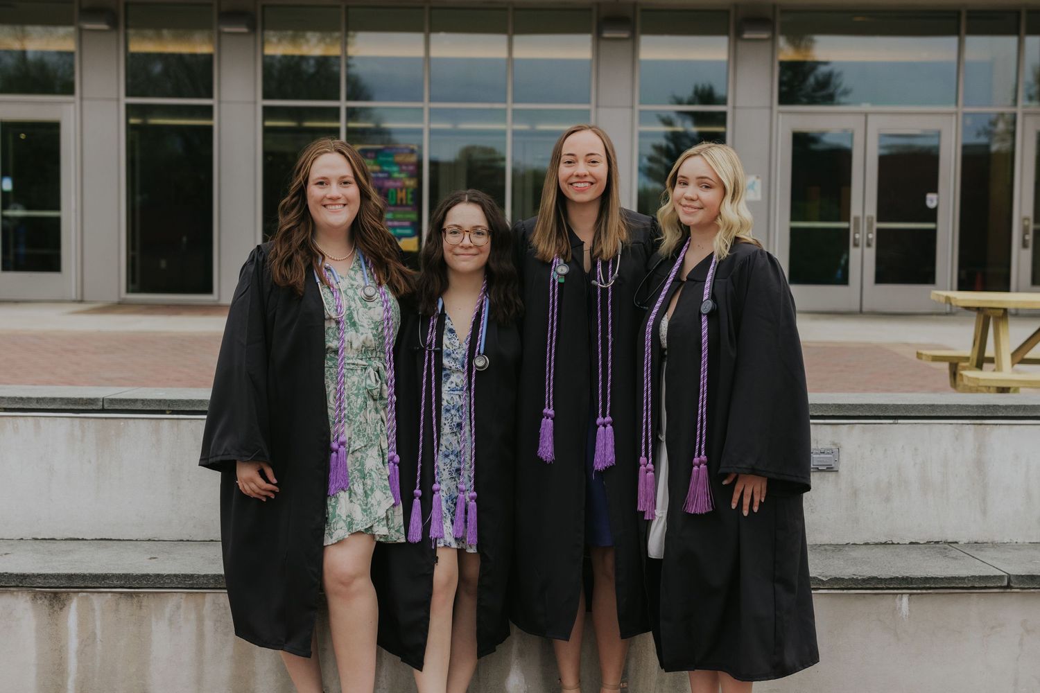 Four nursing graduates in black gowns with purple honor cords posing outside library.