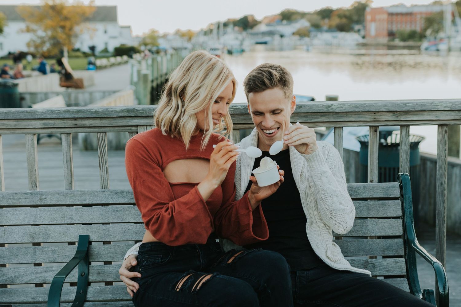 Happy couple sitting on a bench by the waterfront, laughing and enjoying drinks together on a fall day.