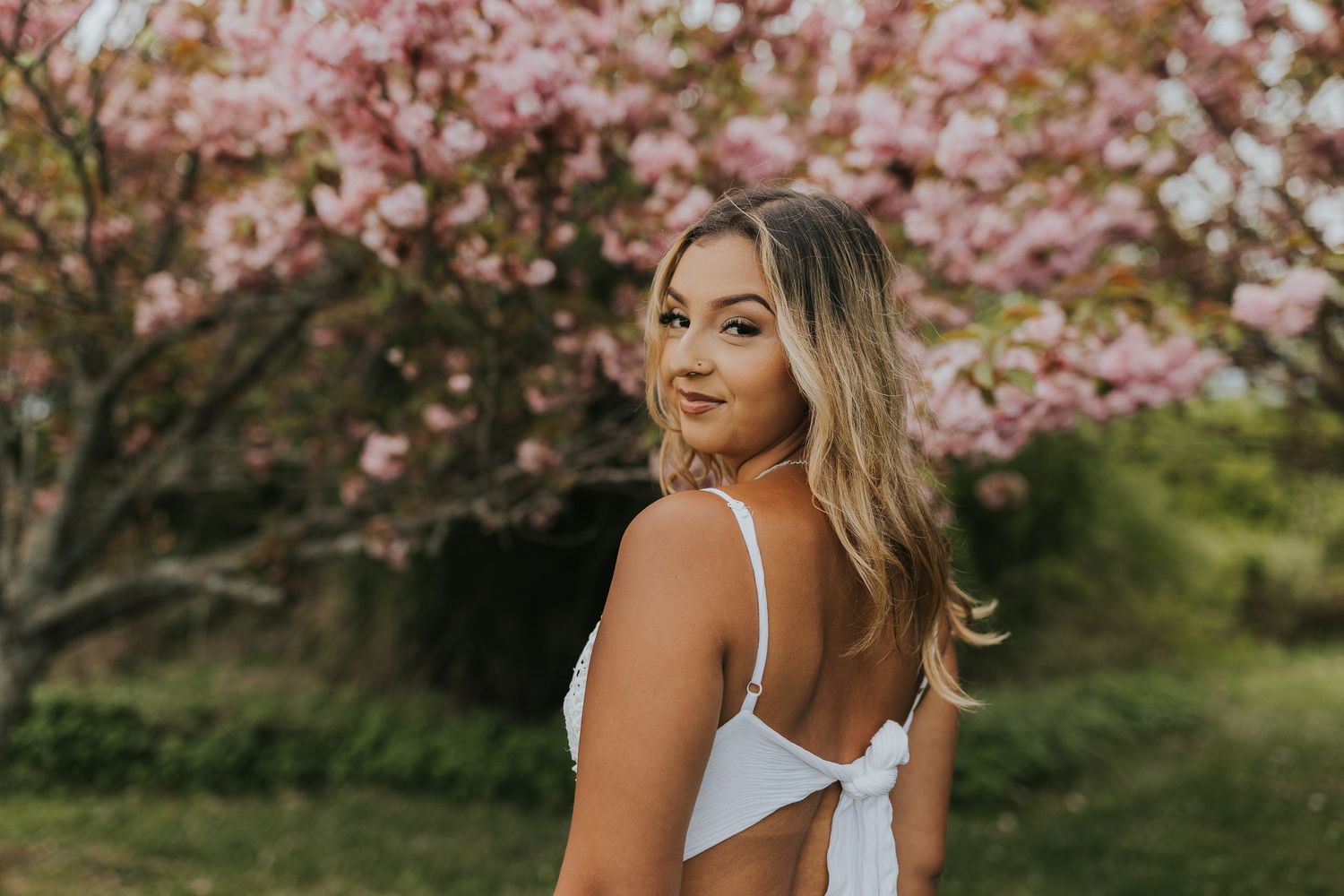 Young woman in white dress posing outdoors near pink cherry blossom trees in spring.