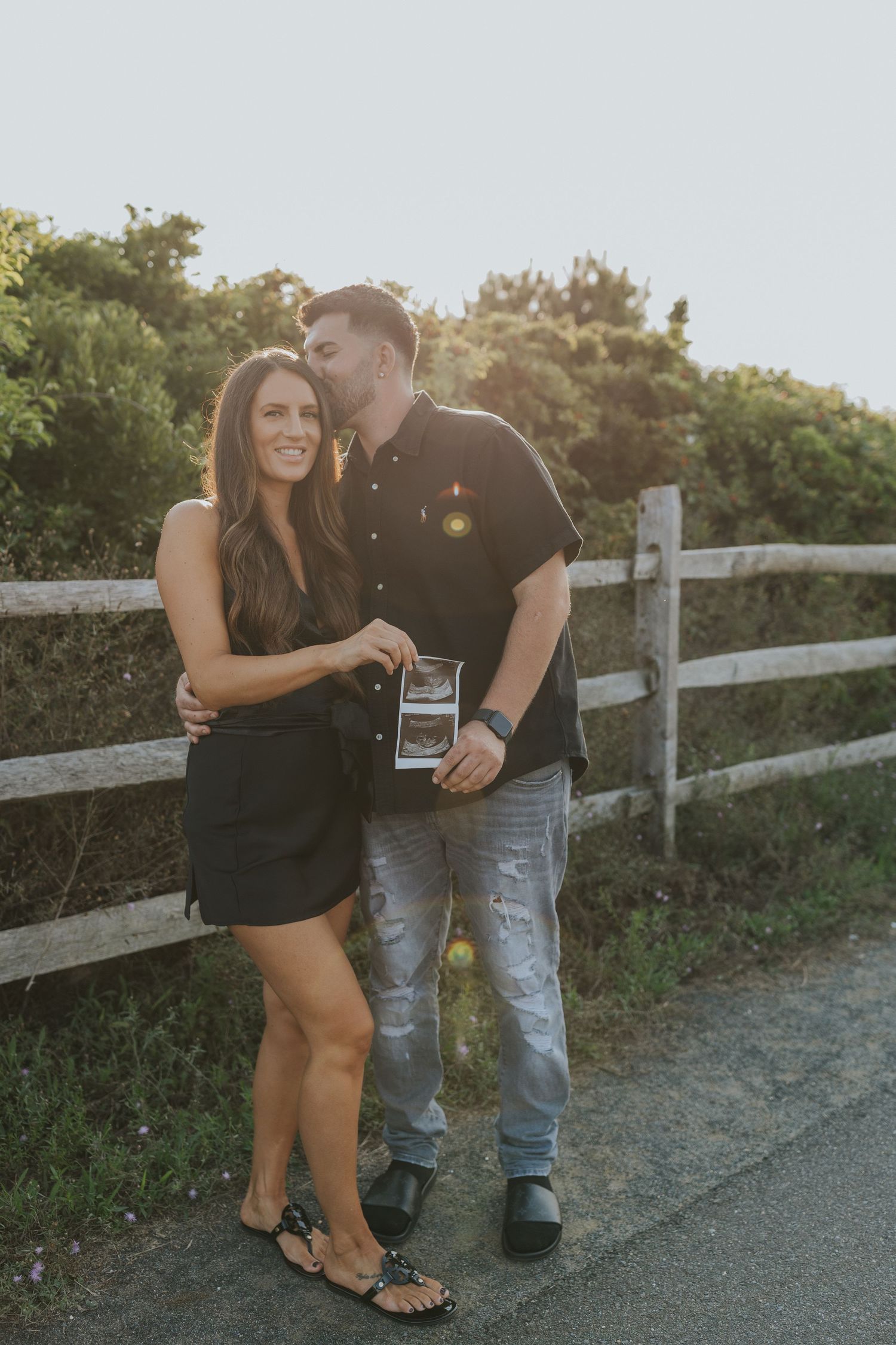 Man kissing woman on cheek while holding ultrasound photo by white fence, pregnancy announcement.