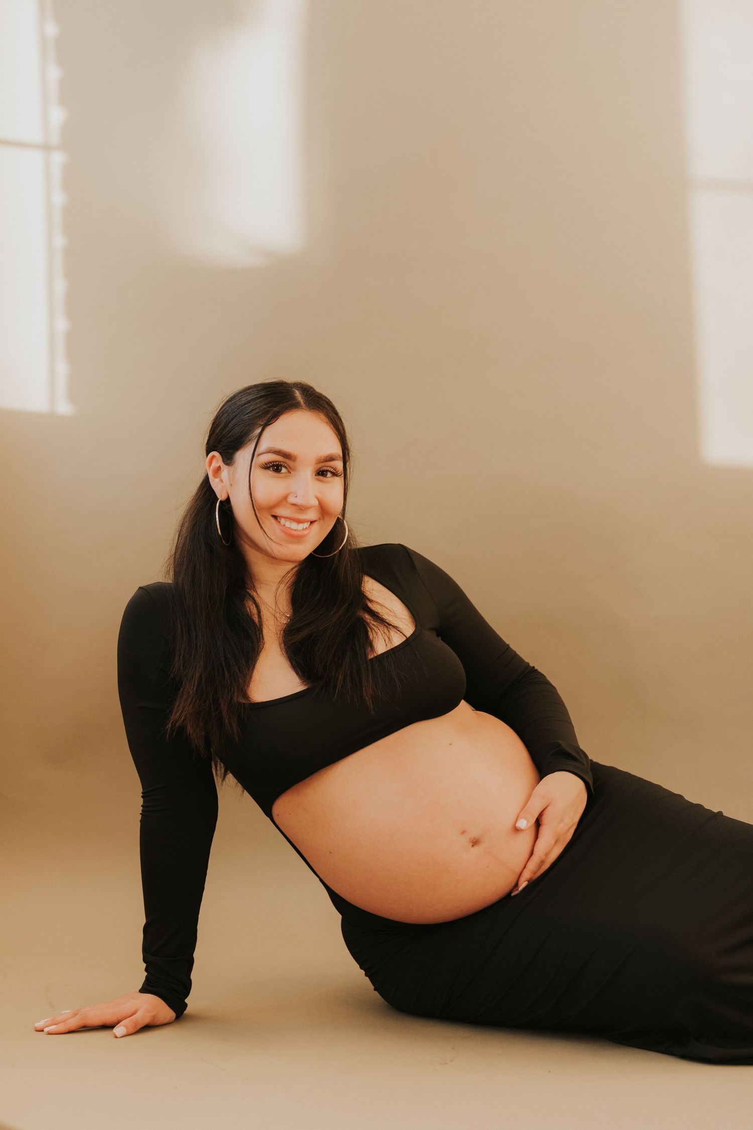 Smiling pregnant woman in black outfit sitting on floor, gently holding her baby bump.
