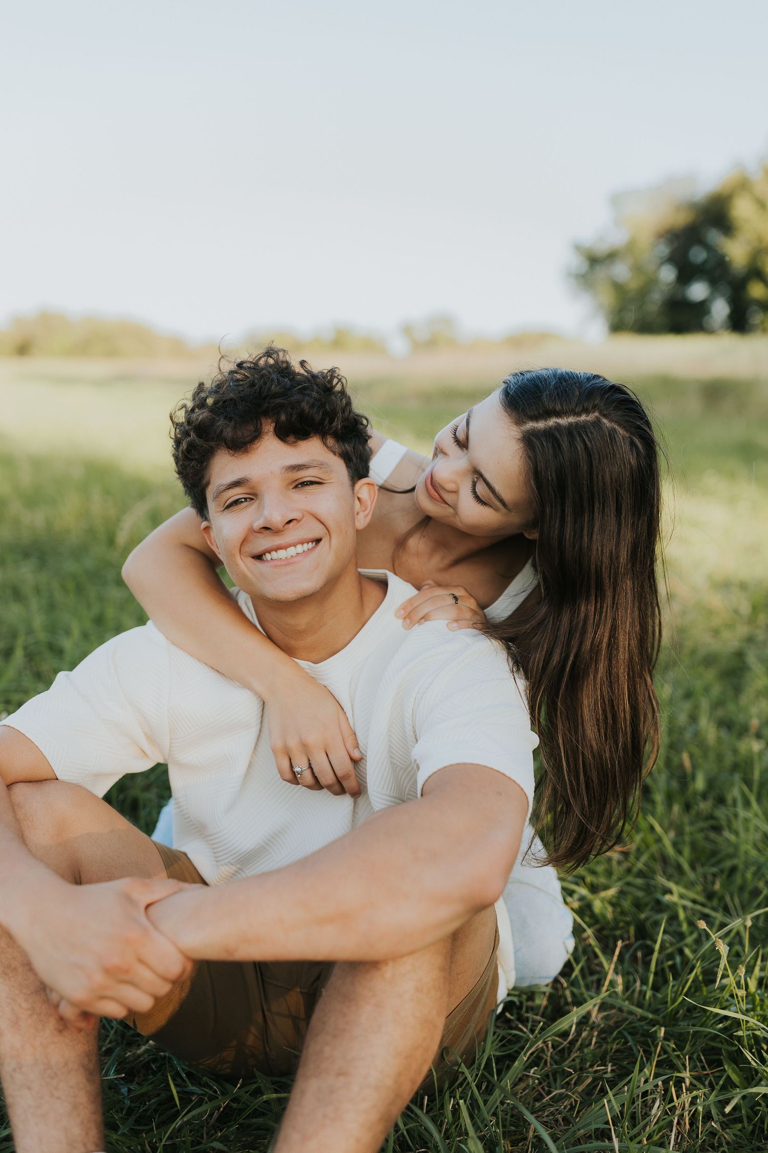 Smiling young man sitting in grass while girlfriend hugs him from behind on sunny day.